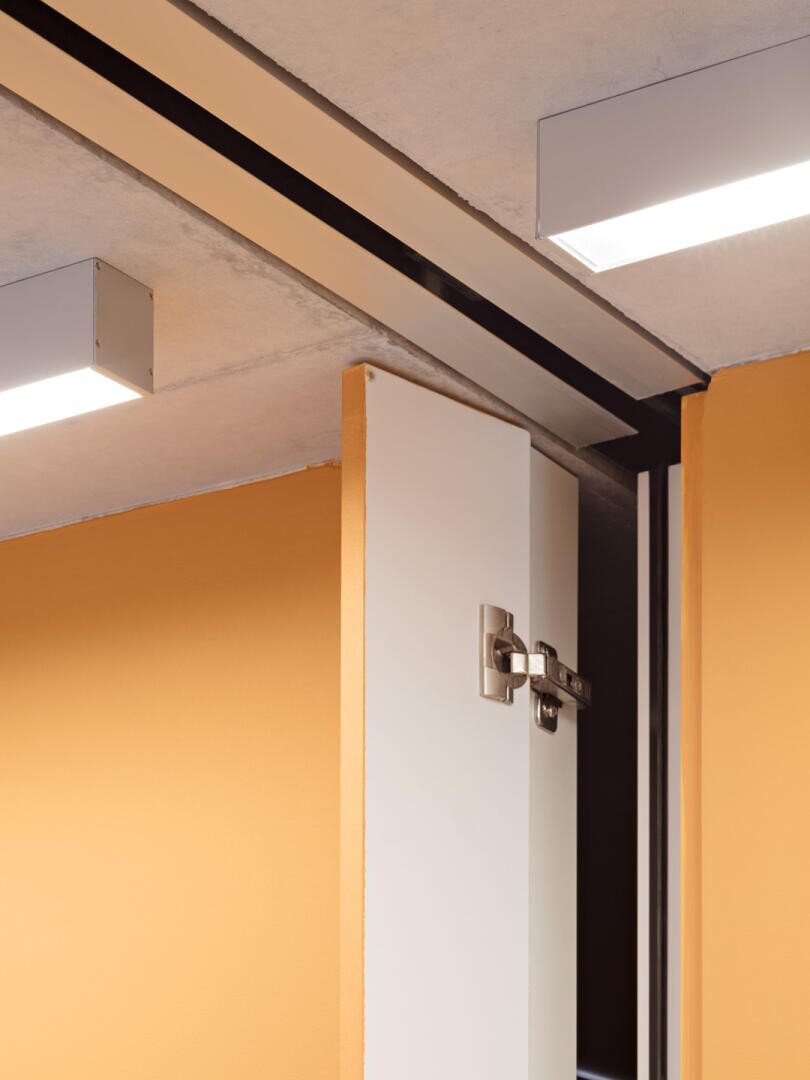 Nahaufnahme einer weißen Tür mit einem Metallscharnier, die teilweise geöffnet ist, vor orangefarbenen Wänden und einer Decke mit rechteckigen Leuchten.
