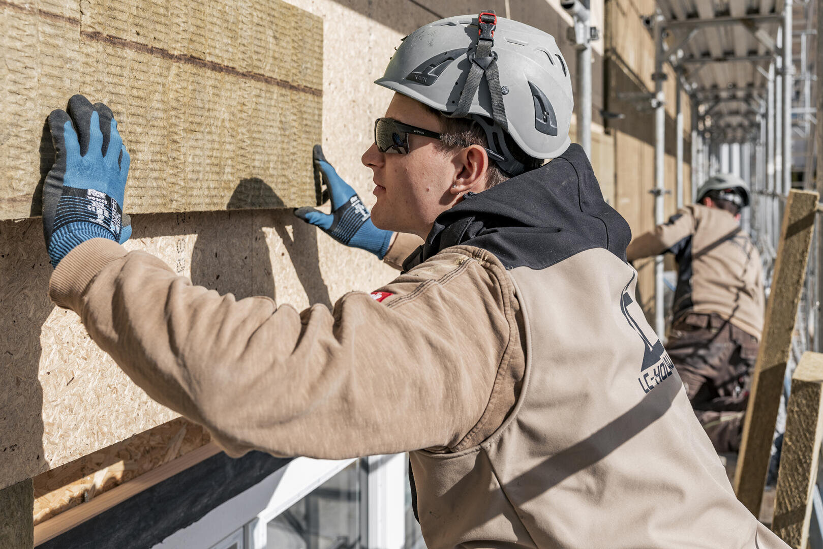 Un ouvrier du bâtiment portant un casque, des gants et des lunettes de sécurité installe des panneaux d'isolation sur le mur extérieur d'un bâtiment, tandis qu'un autre ouvrier effectue un travail similaire à l'arrière-plan.