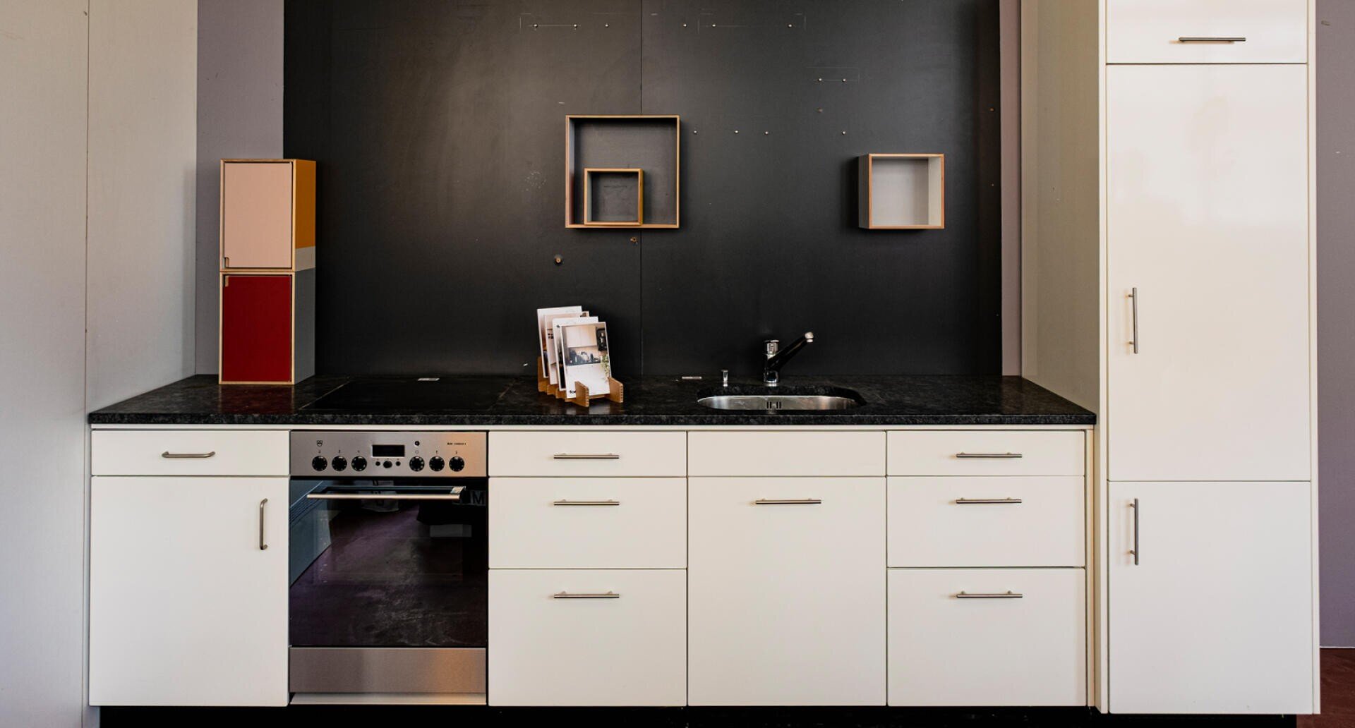 Moderne Küche mit weißen Schränken, schwarzer Arbeitsplatte, Herd mit Backofen, kleiner Spüle und minimalistischem Dekor mit geometrischen Wandregalen und einem kleinen Gerät auf dem Tresen.