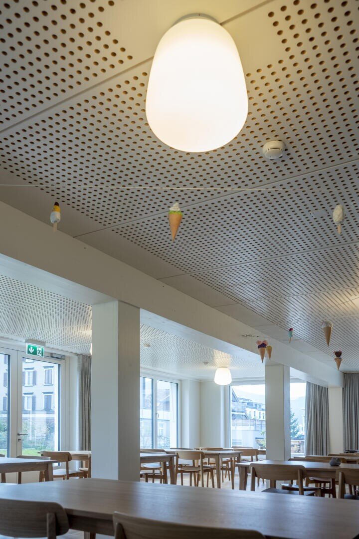 Une cafétéria moderne avec des tables et des chaises en bois, de grandes fenêtres et des plafonds blancs perforés, décorés de petits cornets de glace suspendus. Au premier plan, on peut voir un plafonnier lumineux.