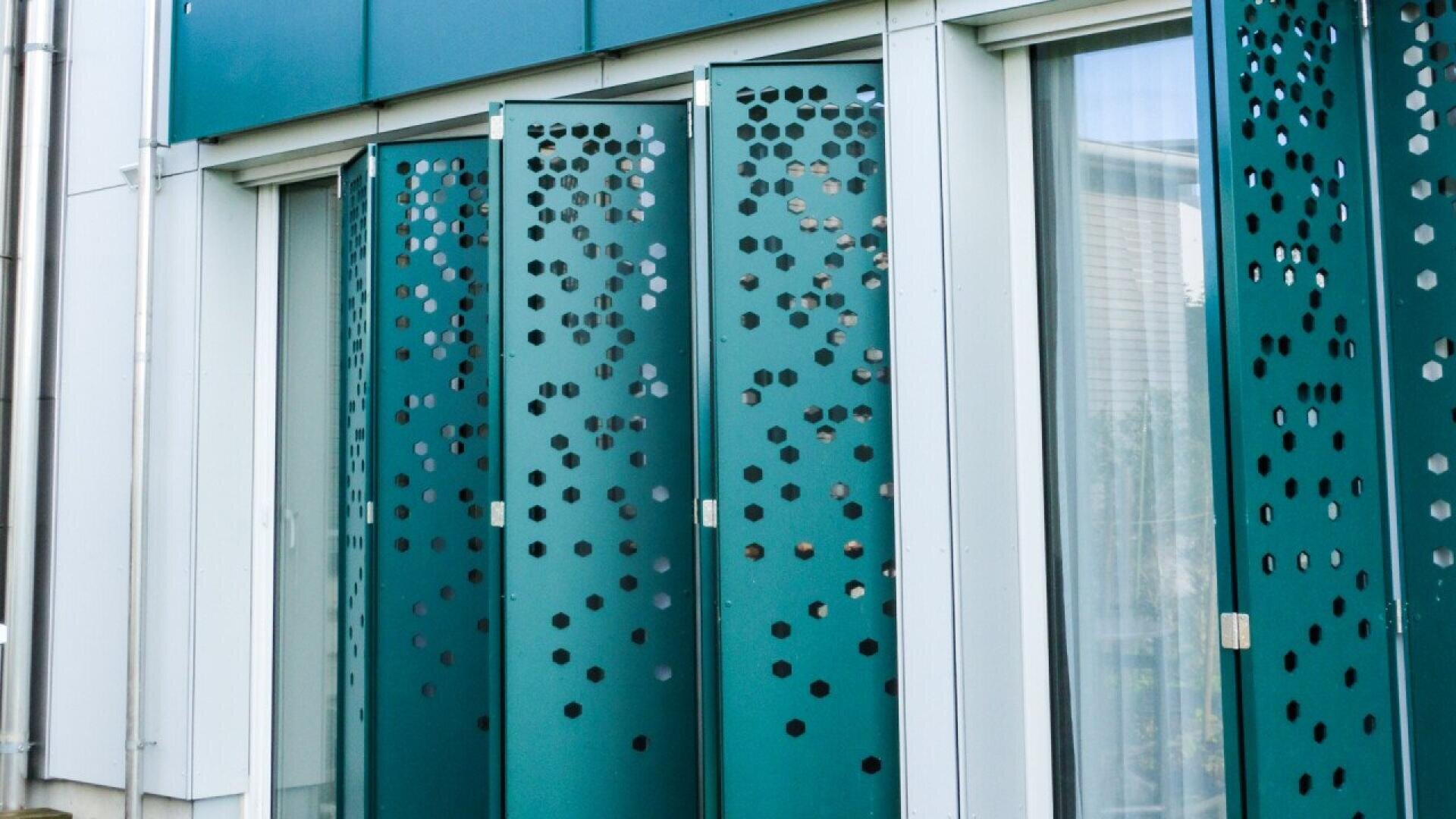 Gros plan sur des panneaux métalliques sarcelle avec des découpes circulaires éparses, recouvrant partiellement les fenêtres d'une façade d'immeuble moderne. Les panneaux créent un effet décoratif et géométrique.