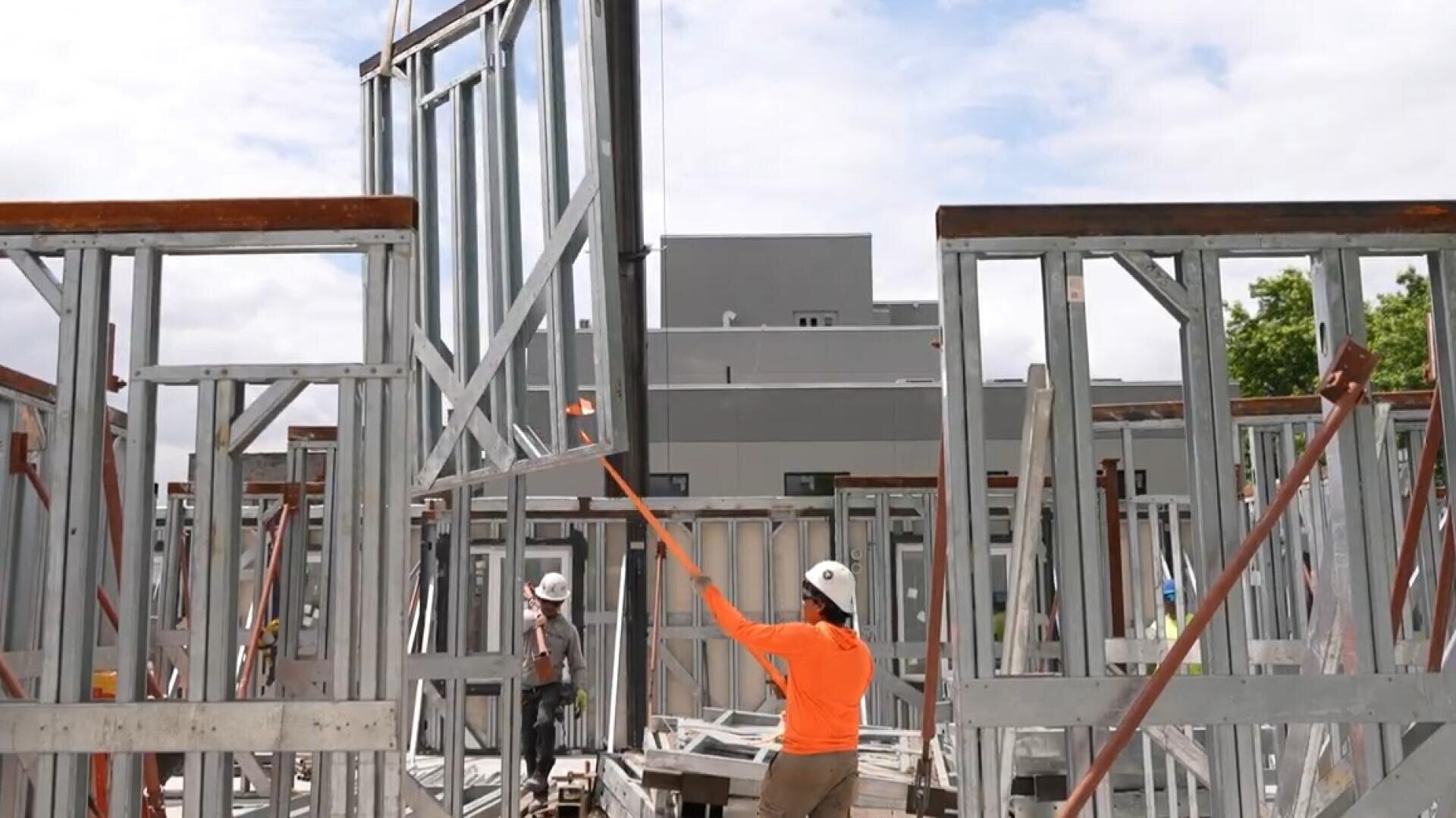 Bauarbeiter, die Schutzhelme tragen, montieren eine Metallrahmenkonstruktion. Ein Arbeiter in einem orangefarbenen Hemd verwendet eine Stange, um eine hohe Metallplatte an ihren Platz zu führen, während ein anderer in der Nähe zwischen den Metallträgern unter einem teilweise bewölkten Himmel steht.