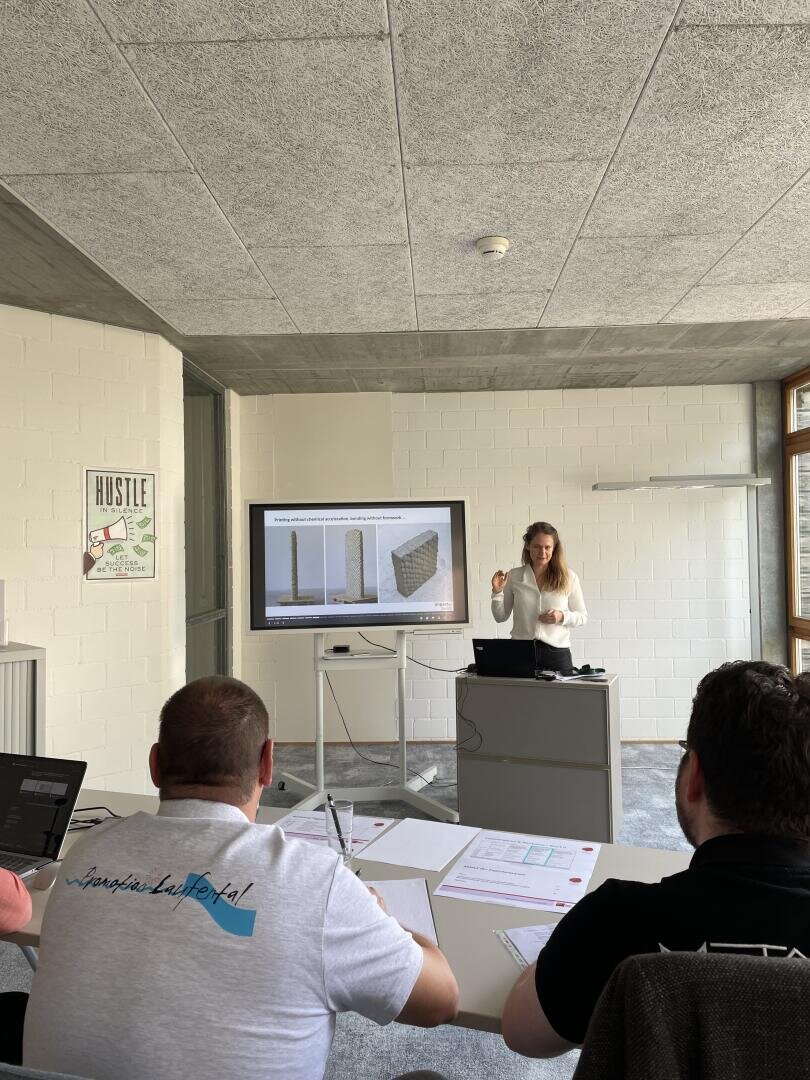 Eine Frau steht vorne in einem Klassenzimmer und hält eine Präsentation mit Bildern von Betonblöcken auf einem Bildschirm, während zwei Männer an Tischen mit Papieren und Laptops sitzen, zuhören und Notizen machen.