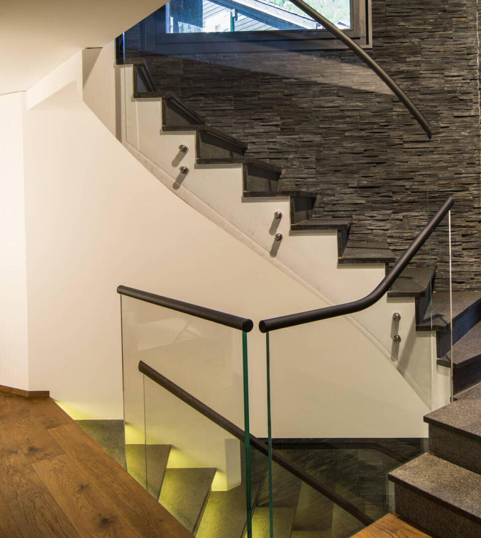Escalier moderne avec rampe en verre et en métal, mur d'accent en pierre et plancher en bois. Un éclairage chaud illumine les marches et le mur inférieur, créant un intérieur moderne et élégant.