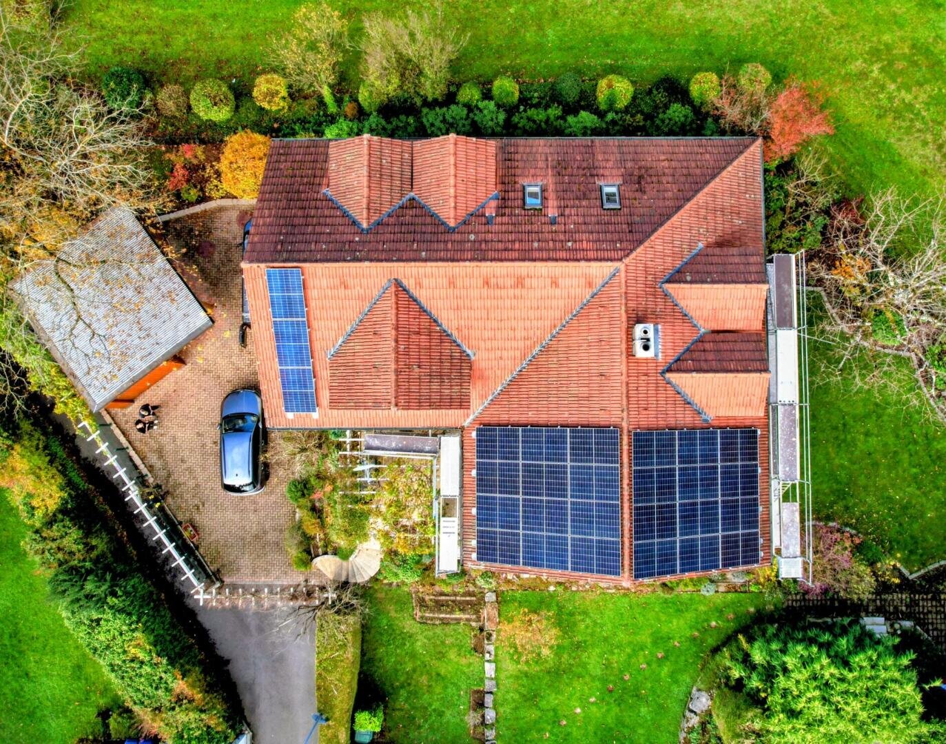 Luftaufnahme eines Hauses mit rotem Ziegeldach, das mit Sonnenkollektoren ausgestattet ist, einer Einfahrt mit geparktem Auto, einem kleinen Schuppen und grünem Rasen, umgeben von Bäumen und Sträuchern in einem angelegten Garten.