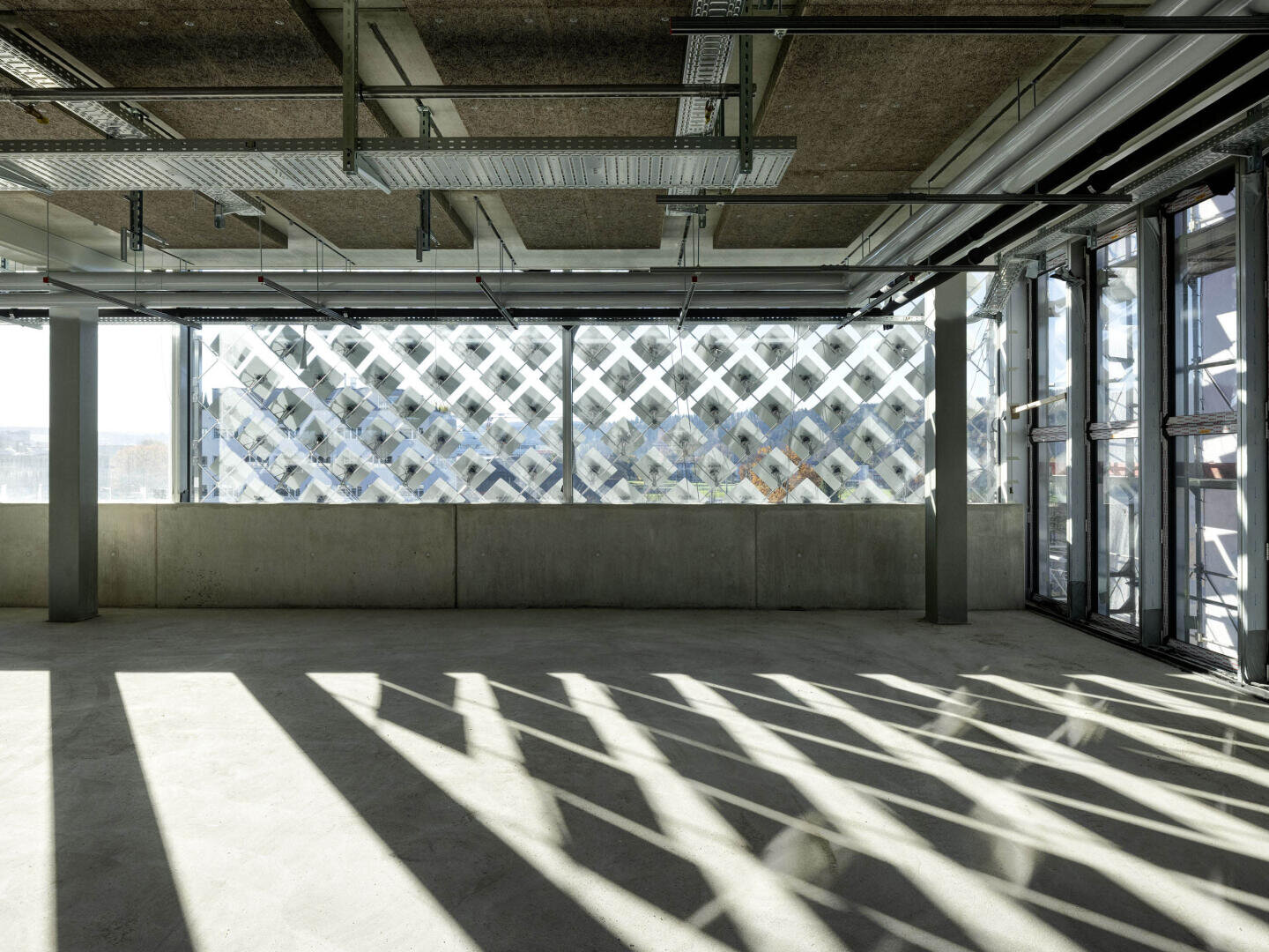 Un espace industriel vide avec des sols en béton, des plafonds apparents, de grandes fenêtres et la lumière du soleil qui projette des ombres géométriques sur le sol à partir d'une structure extérieure à motifs.