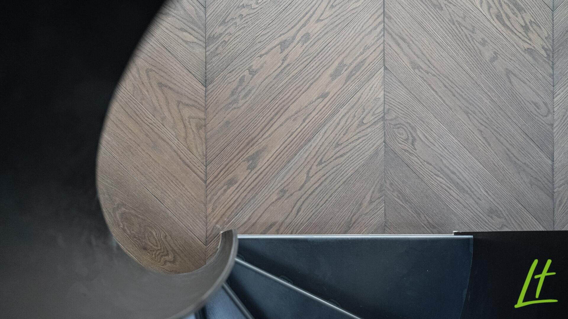 Draufsicht auf eine Wendeltreppe mit dunklen Stufen und einem Holzboden mit Chevron-Muster. In der unteren rechten Ecke ist ein grünes lt-Logo zu sehen.