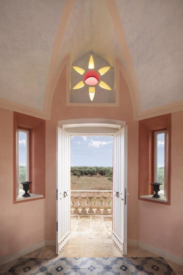 Une paire de doubles portes blanches s'ouvre sur un balcon donnant sur un champ et des arbres. Au-dessus des portes se trouve une fenêtre en forme de soleil, et deux petites fenêtres avec des vases noirs encadrent les portes. Les murs de la pièce sont roses et le sol est recouvert d'un carrelage à motifs.