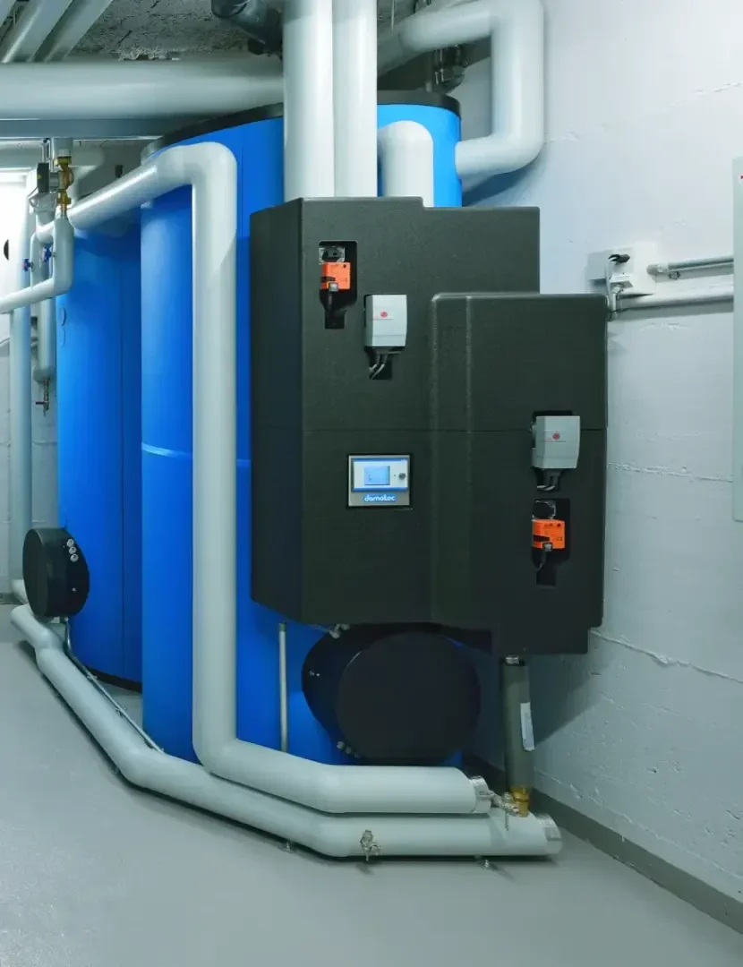 Große blaue Wassertanks sind mit weißen isolierten Rohren und schwarzen Schalttafeln in einem sauberen, modernen Service- oder Heizraum mit grauem Boden und weiß gestrichenen Wänden verbunden.