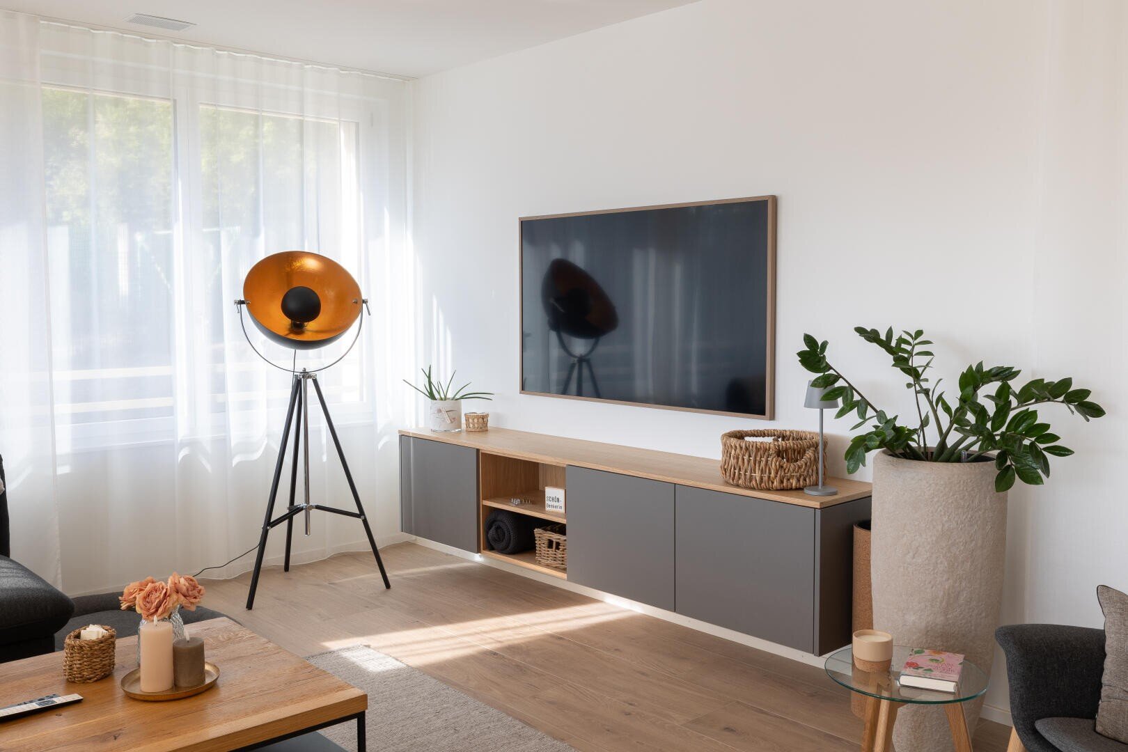 Ein modernes Wohnzimmer mit einem Fernseher an der Wand, einer großen Stehlampe, Topfpflanzen, einem hölzernen Couchtisch mit Blumen und Büchern und einem Fenster mit durchsichtigen weißen Vorhängen, die natürliches Licht hereinlassen.