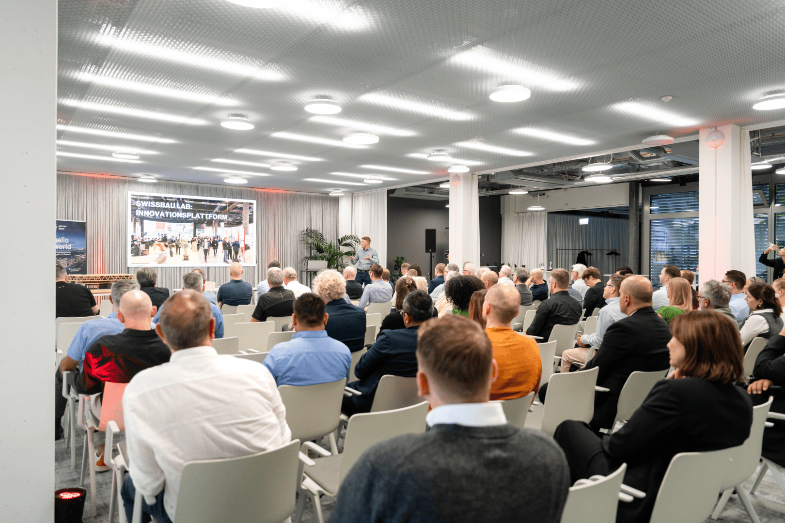 Eine Gruppe von Personen sitzt in einem modernen Konferenzraum und blickt auf einen großen Bildschirm, auf dem eine Präsentation gezeigt wird. Der Raum ist mit weißen Stühlen und Deckenleuchten gut beleuchtet, und einige Personen stehen in der Nähe der Bühne.
