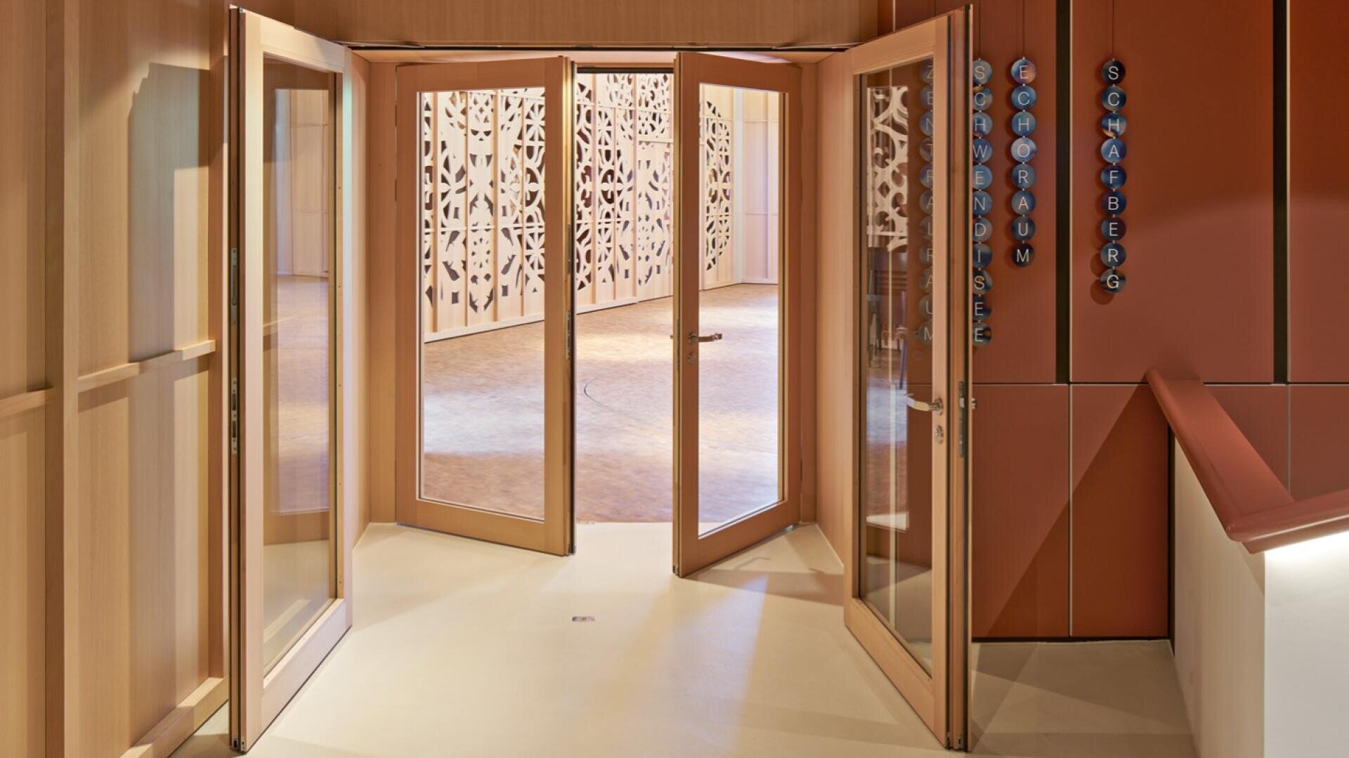 Vier gläserne Doppeltüren führen in einen geräumigen, gut beleuchteten Raum mit dekorativen, gemusterten Wandpaneelen. Das Ambiente ist in warmen Holz- und Neutraltönen gehalten und schafft eine einladende Atmosphäre.