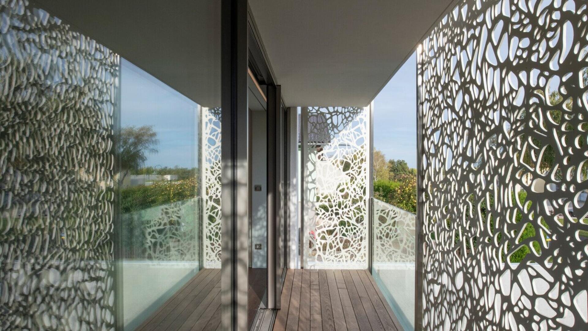 Un balcon moderne avec un sol en bois et des panneaux décoratifs en métal blanc présentant un motif de découpe organique et abstrait, qui projette des ombres complexes sur les murs et le sol. Des portes vitrées et de la verdure sont visibles à l'extérieur.