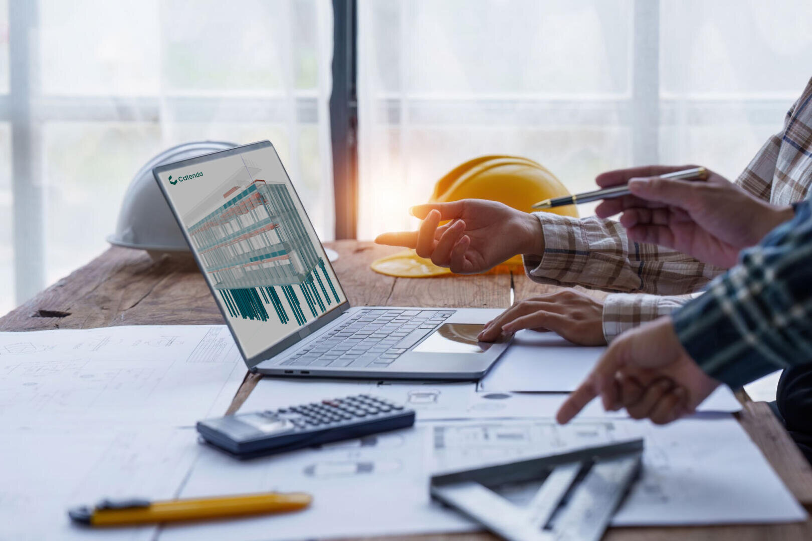 Deux personnes discutent de données affichées sur un ordinateur portable à une table en bois avec des plans de construction, une calculatrice, une règle, des crayons et des casques de protection, ce qui laisse supposer une discussion sur un projet de construction ou d'ingénierie.