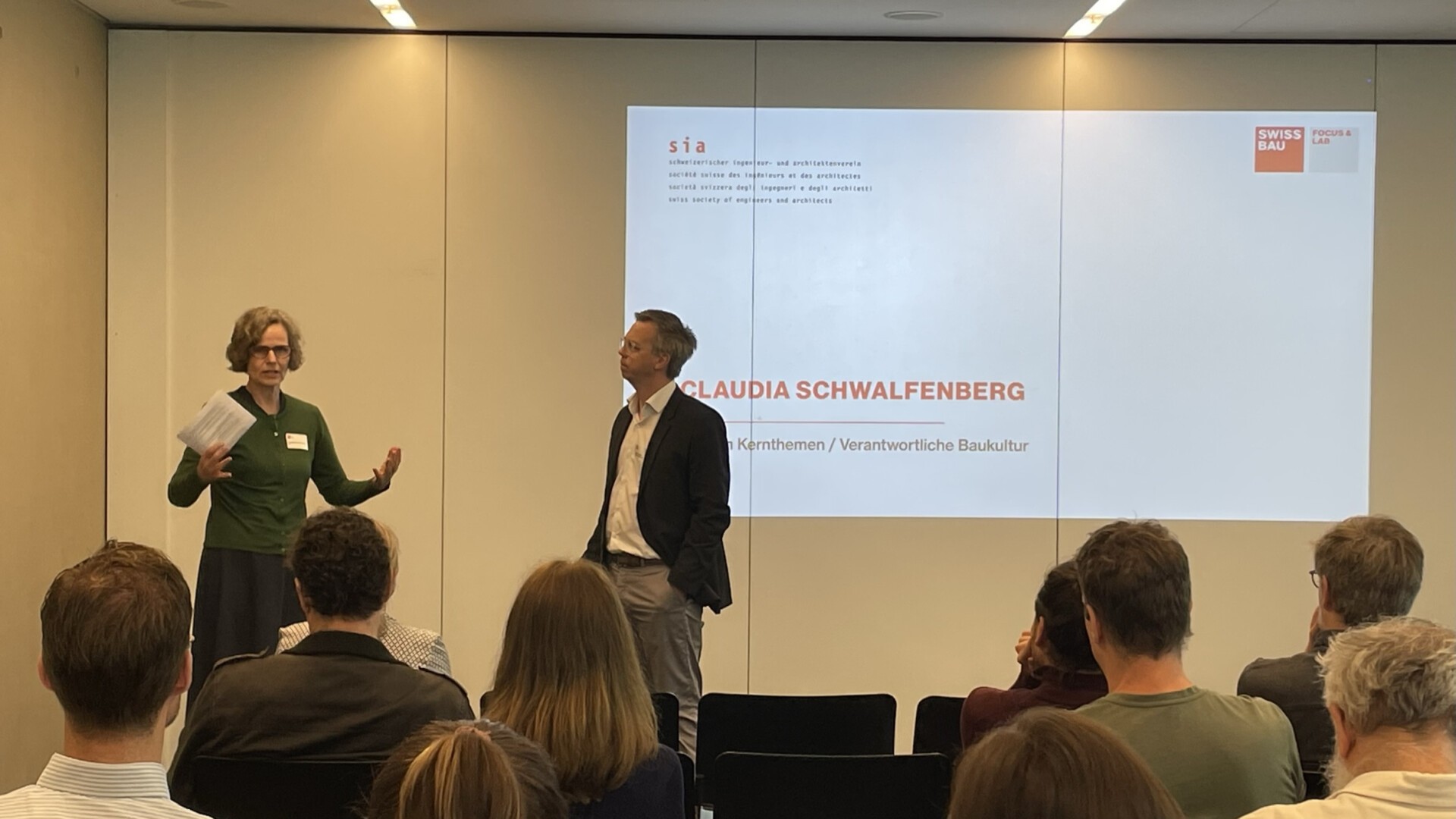 Zwei Personen stehen vor einem sitzenden Publikum und präsentieren. Die Frau links gestikuliert mit Notizen; der Mann rechts hört zu. Hinter ihnen ist ein Bildschirm mit dem Namen „Claudia Schwalfenberg“ und dem Swissbau-Branding zu sehen.