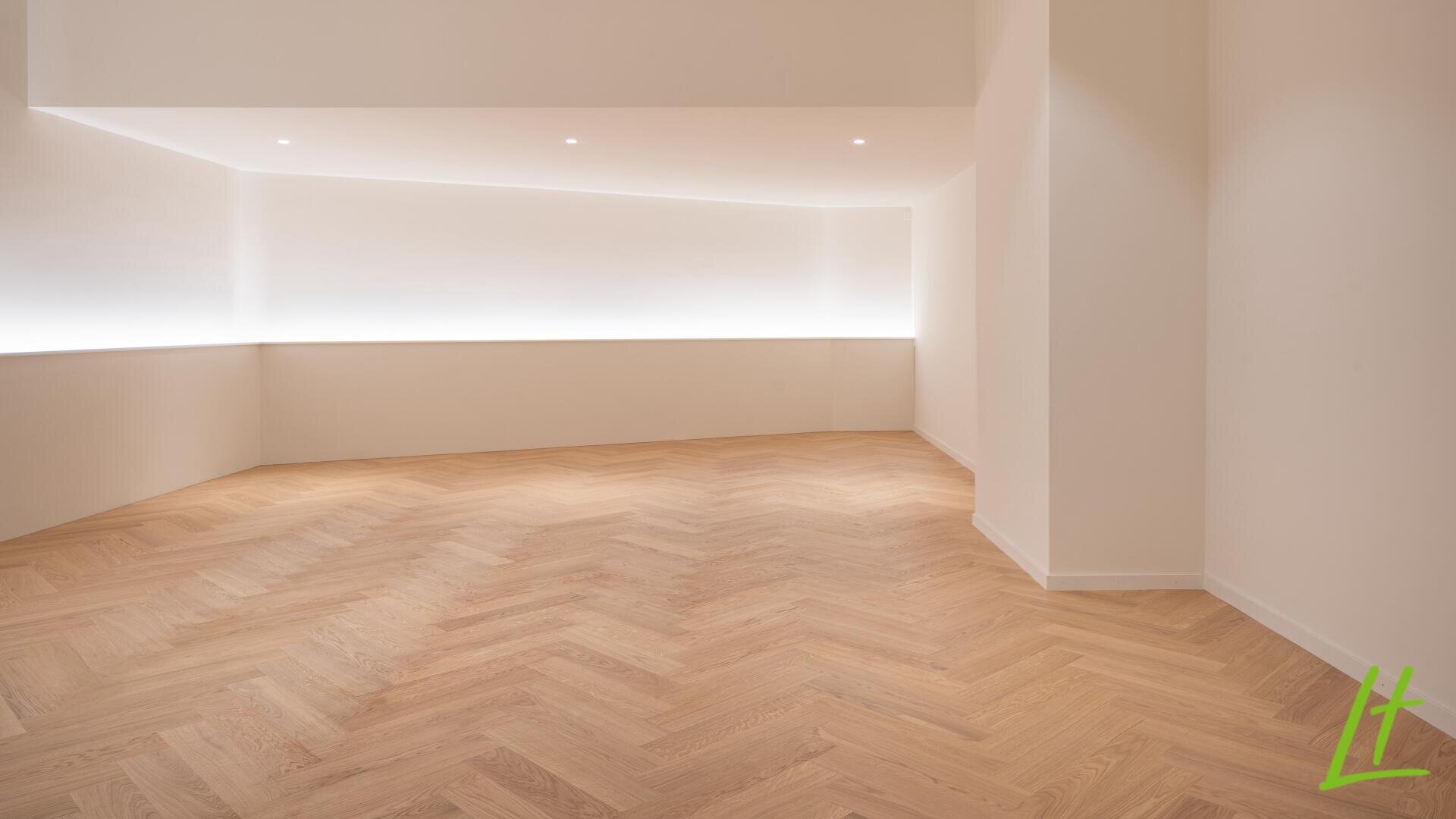Une pièce moderne et vide avec un sol en bois à chevrons, des murs blancs et des éclairages intégrés le long du plafond et des murs, créant un espace lumineux et minimaliste.