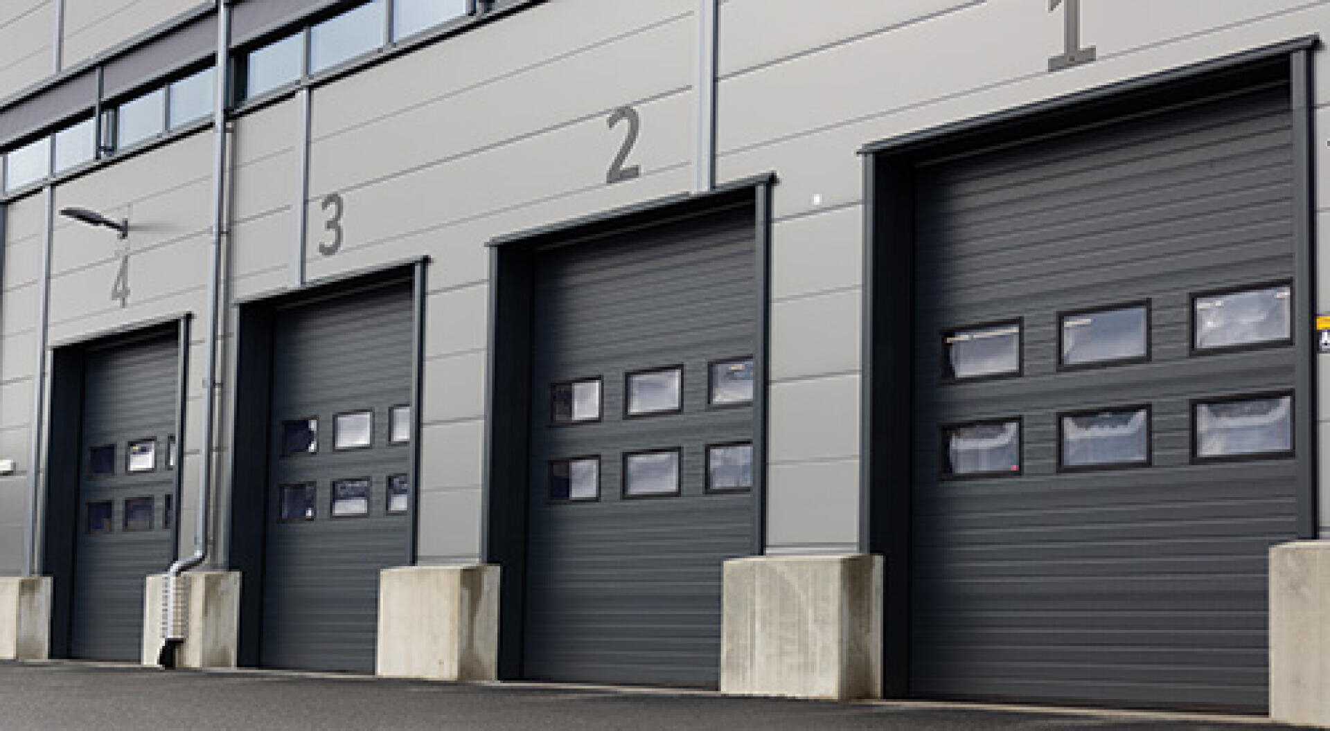 Quatre portes de garage industrielles avec de petites fenêtres et numérotées de 1 à 4 se trouvent dans un bâtiment gris moderne. Des barrières en béton sont fixées à la base de chaque porte.