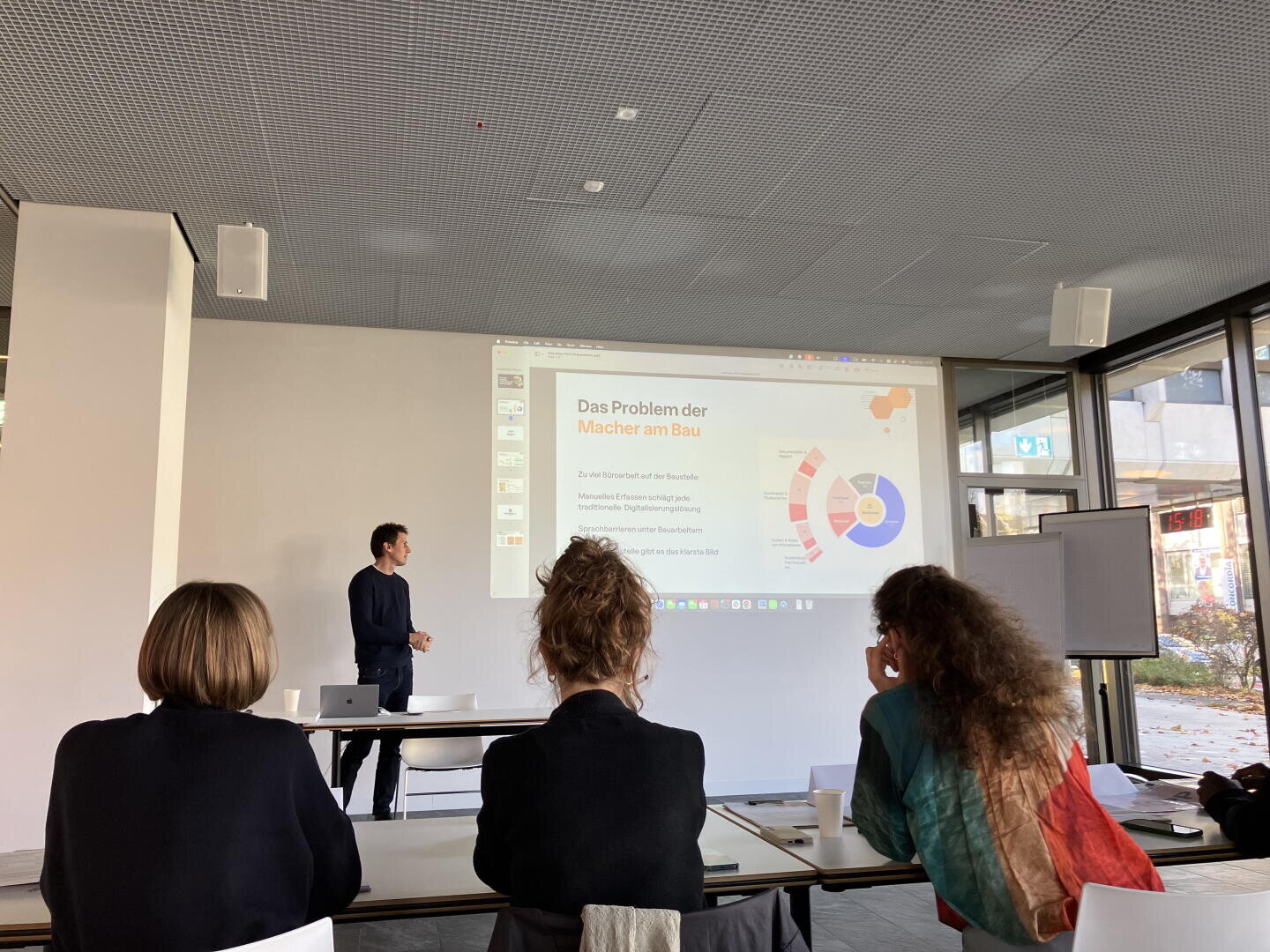 Ein Mann hält einen Vortrag in einem modernen, gut beleuchteten Raum vor vier sitzenden Personen. Ein Dia in deutscher Sprache mit Text und einer bunten Grafik wird an die Wand hinter ihm projiziert. Große Fenster zeigen eine städtische Szene draußen.