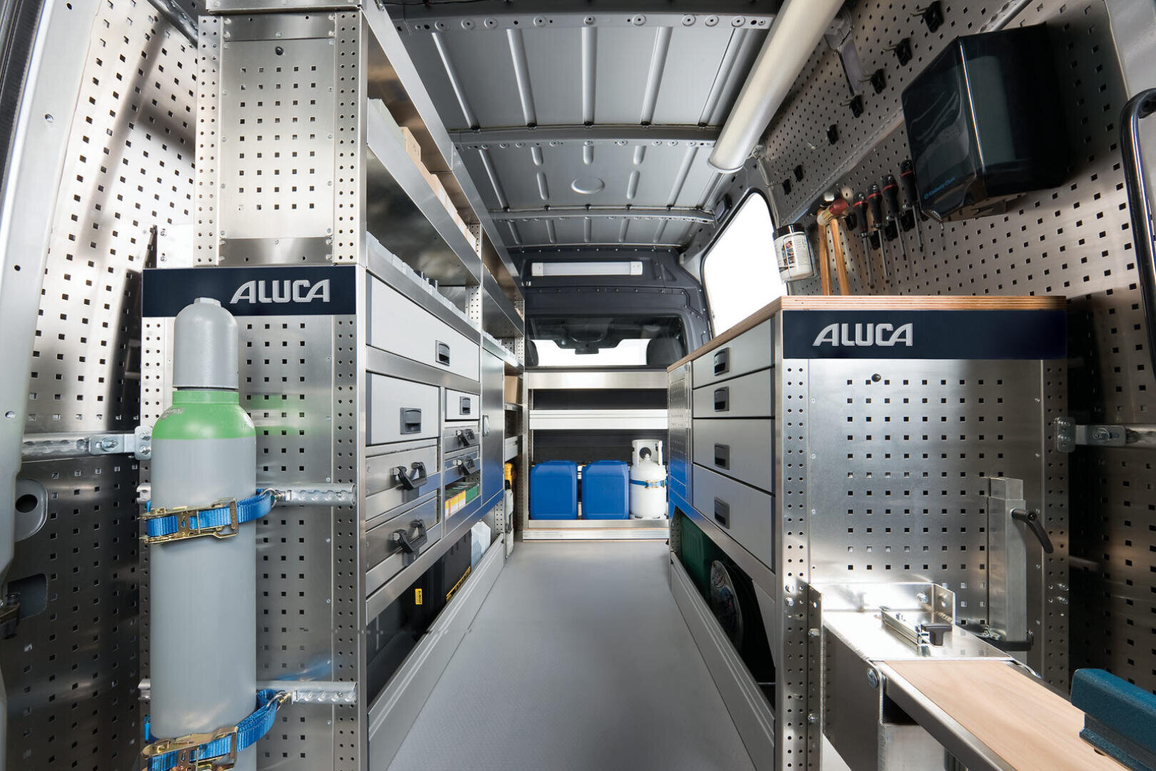 Das Innere eines gut organisierten Arbeitswagens mit Metallregalen, Schubladen und Werkzeugen, die ordentlich auf beiden Seiten angeordnet sind. Auf der linken Seite ist eine Gasflasche gesichert, und im hinteren Bereich sind verschiedene Behälter und Kisten zu sehen.