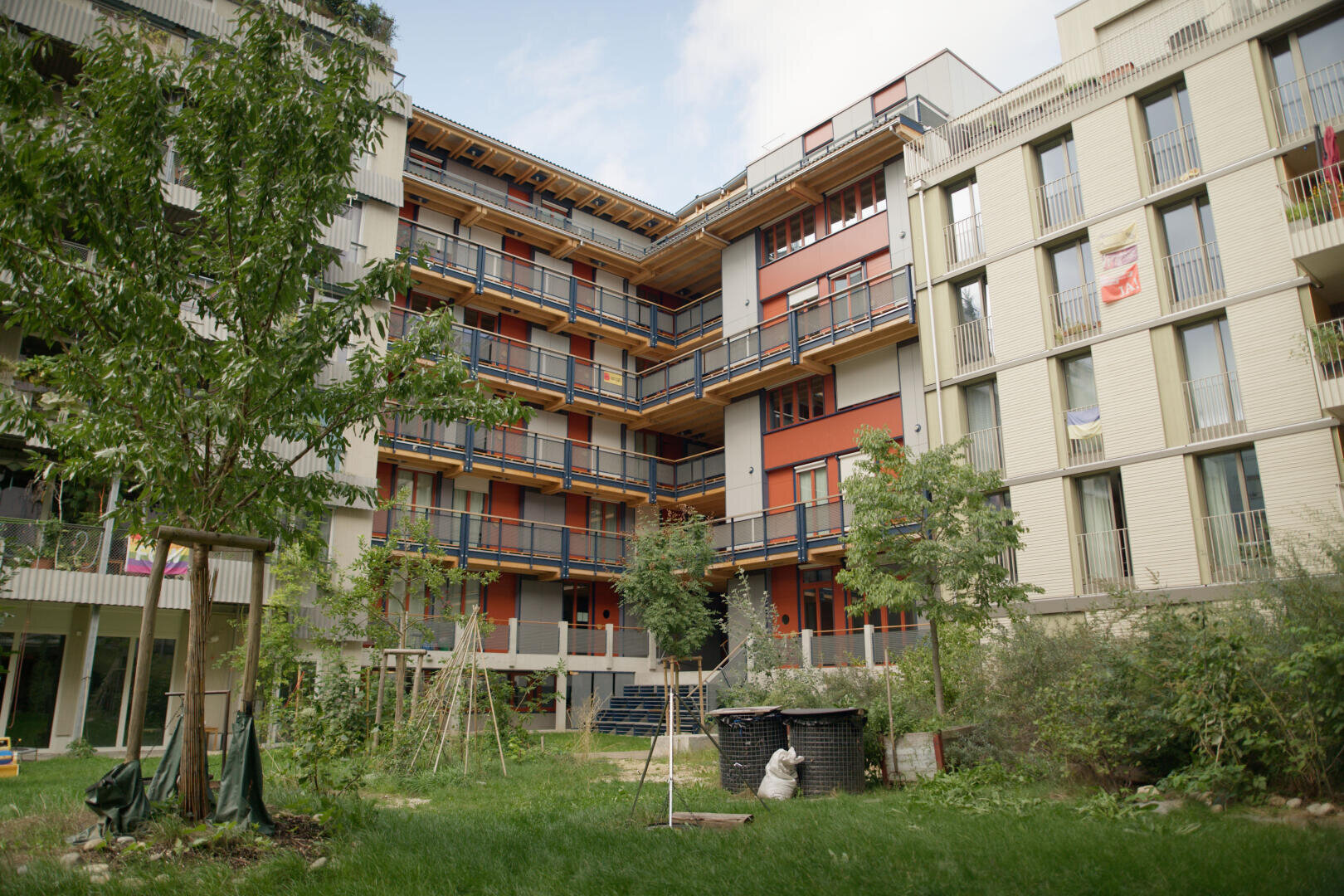 Ein modernes Mehrfamilienhaus mit Balkonen blickt auf einen grünen Innenhof mit Bäumen, Pflanzen und einem kleinen runden Gebäude in der Mitte. Der Himmel ist teilweise bewölkt.