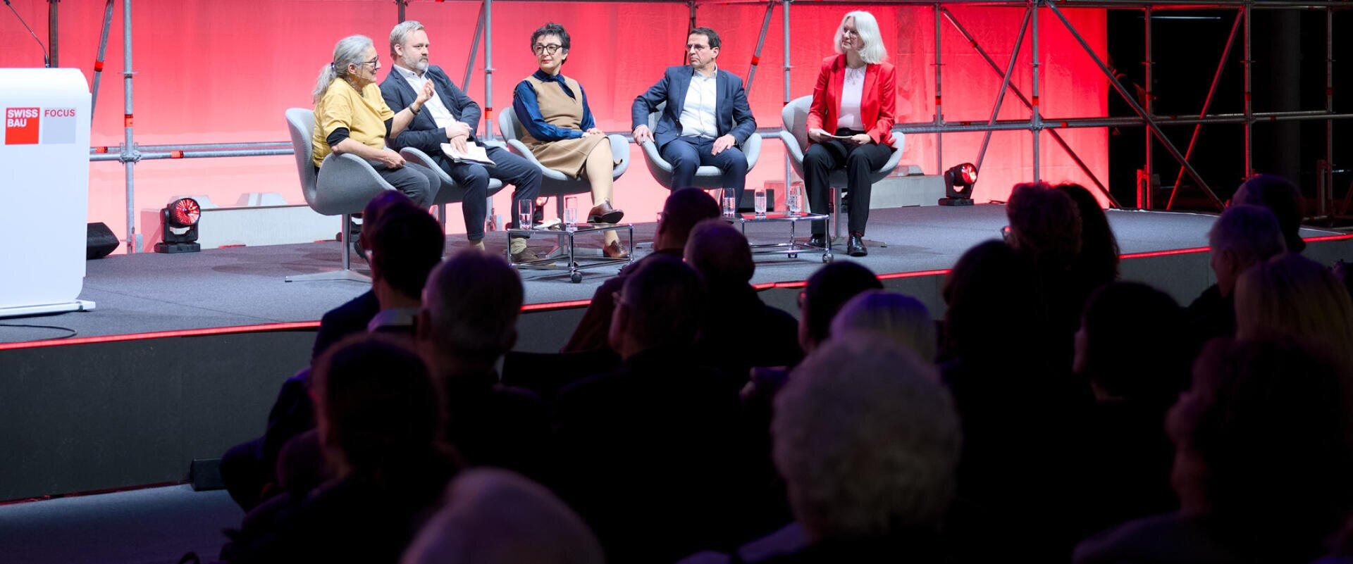 Fünf Personen sitzen bei einer Podiumsdiskussion auf der Bühne und blicken auf das Publikum. Die Bühne ist mit roten und weißen Lichtern beleuchtet, und links befindet sich ein Podium. Im Hintergrund ist ein Metallgerüst zu sehen.