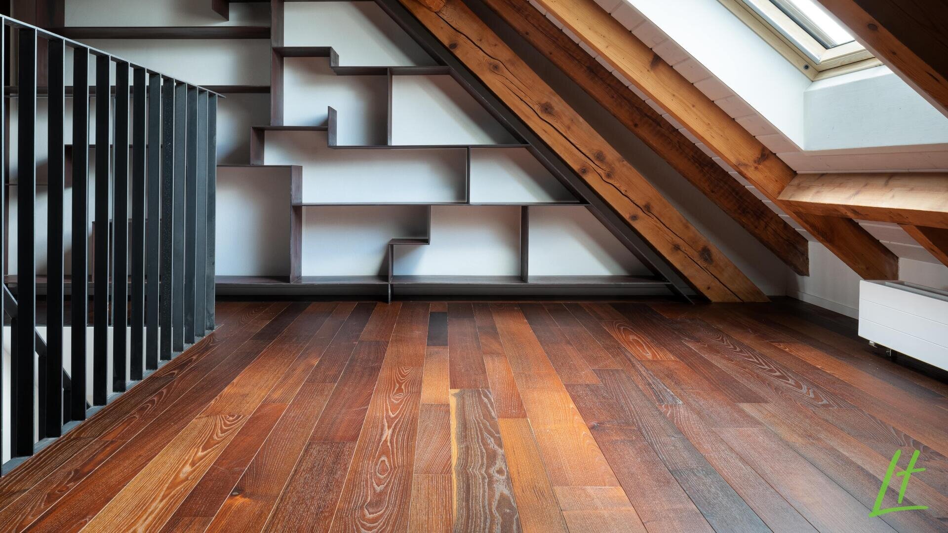 Ein modernes Dachzimmer mit schräger Holzdecke, Oberlicht, dunklem Holzboden, Metallgeländer auf der linken Seite und einem geometrischen Bücherregal an der Rückwand.