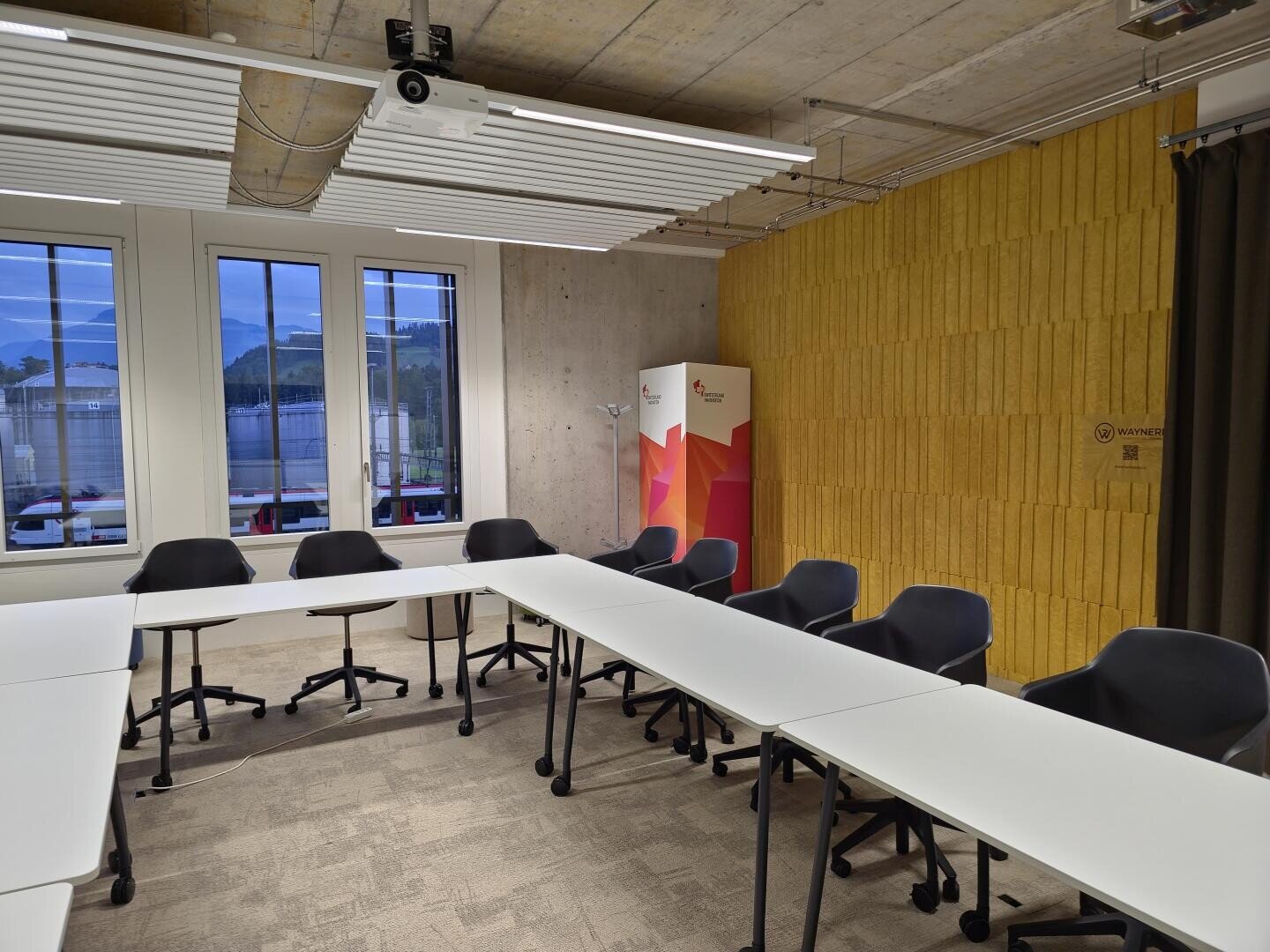 Ein moderner Konferenzraum mit U-förmigen weißen Tischen, schwarzen Stühlen, großen Fenstern mit Blick nach draußen, einem Projektor an der Decke und einem farbenfrohen Schrank vor einer gelb strukturierten Wand.
