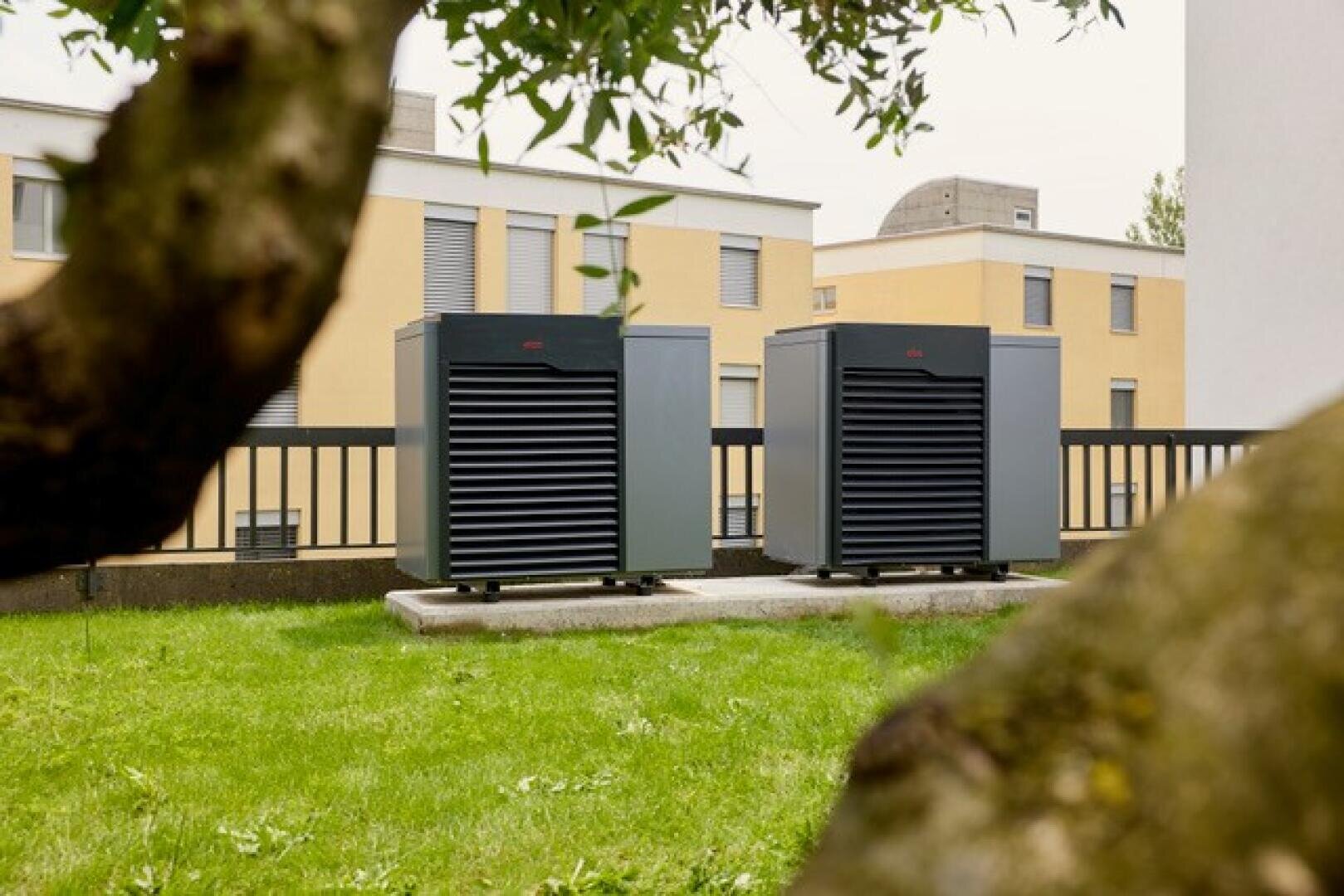 Zwei große Wärmepumpenaggregate für den Außenbereich stehen auf Betonplatten auf einer Wiese, mit modernen gelben Wohngebäuden und einem schwarzen Zaun im Hintergrund. Baumzweige rahmen die Szene im Vordergrund teilweise ein.