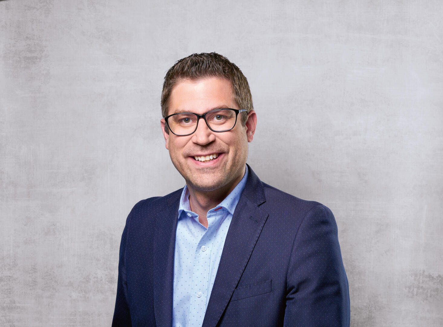 Ein lächelnder Mann mit kurzen braunen Haaren und Brille, der eine blaue Anzugsjacke und ein hellblaues Hemd trägt, steht vor einem hellgrauen, strukturierten Hintergrund.