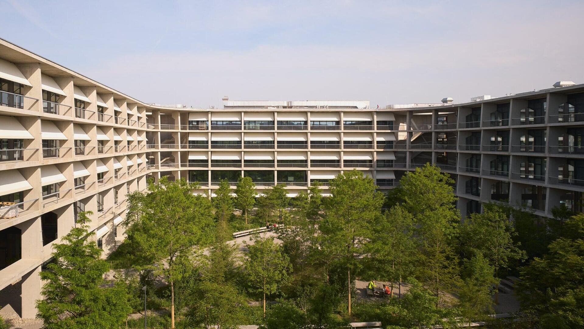 Ein großes, modernes Gebäude mit mehreren Stockwerken umgibt einen zentralen Hof mit Bäumen und Grünpflanzen unter einem teilweise bewölkten Himmel.