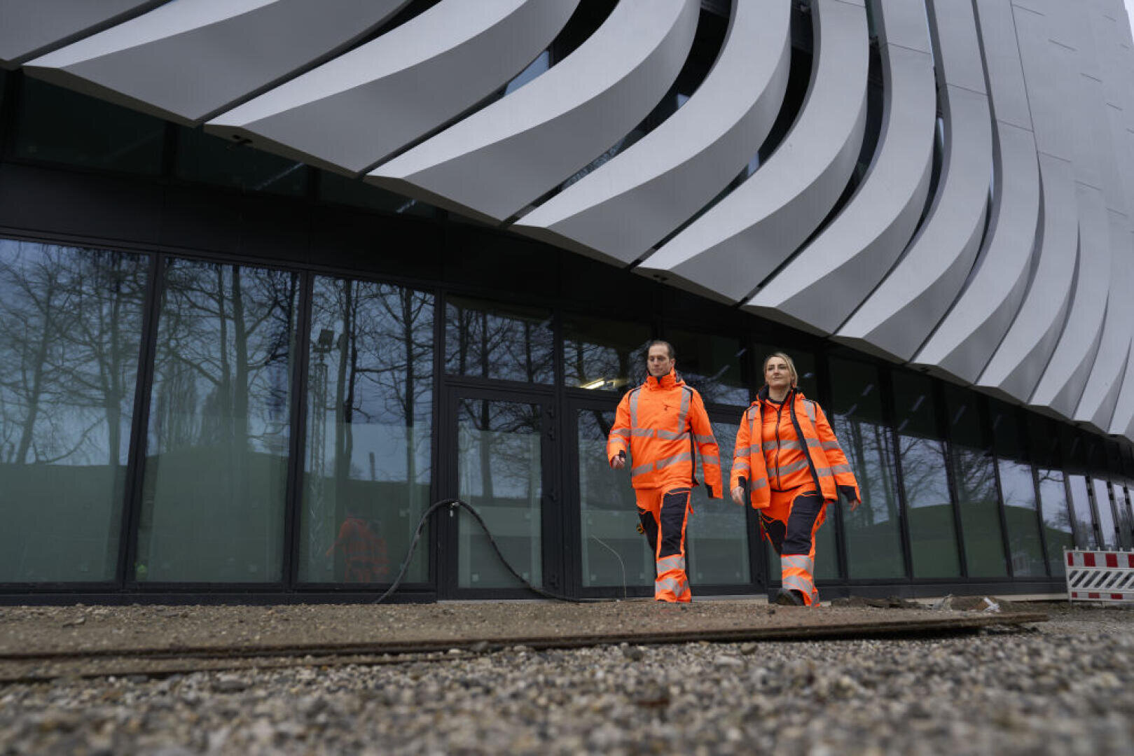 Deux personnes vêtues de gilets de sécurité orange vif et de pantalons sombres marchent côte à côte sur un chemin de gravier devant un bâtiment moderne à l'architecture métallique et incurvée.