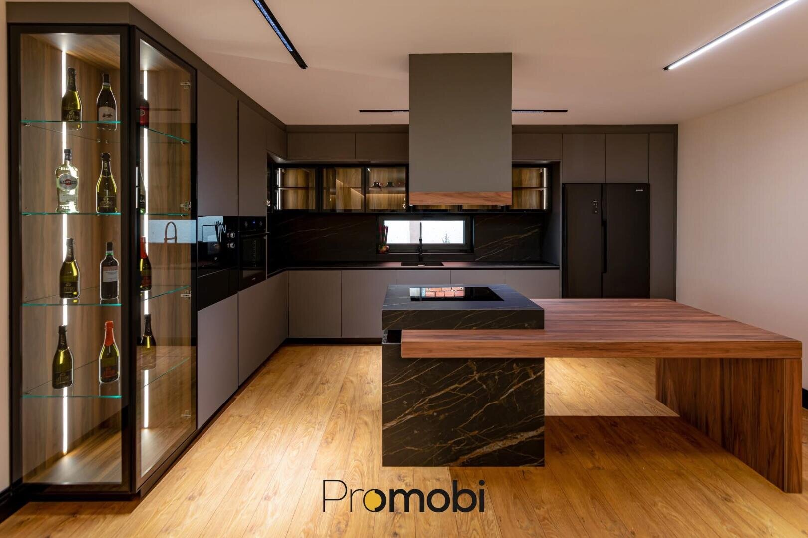 Moderne Küche mit eleganten Schränken, einer Flaschenvitrine, einer schwarzen Insel mit einer Thekenverlängerung aus Holz, Einbaugeräten, einem schwarzen Kühlschrank und einem Holzboden. Das Wort Promobi erscheint unten in der Mitte.