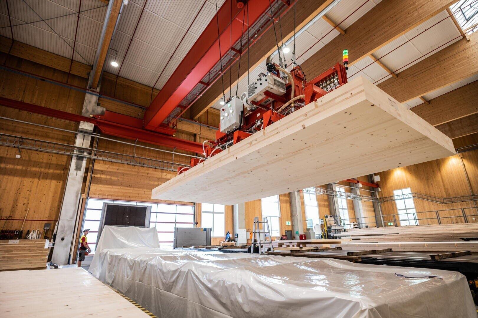 In einer geräumigen, modernen Fabrik mit holzgetäfelten Wänden und hohen Decken wird eine große Holzplatte von einem roten Brückenkran angehoben. Im Hintergrund sind Arbeiter und Maschinen zu sehen.