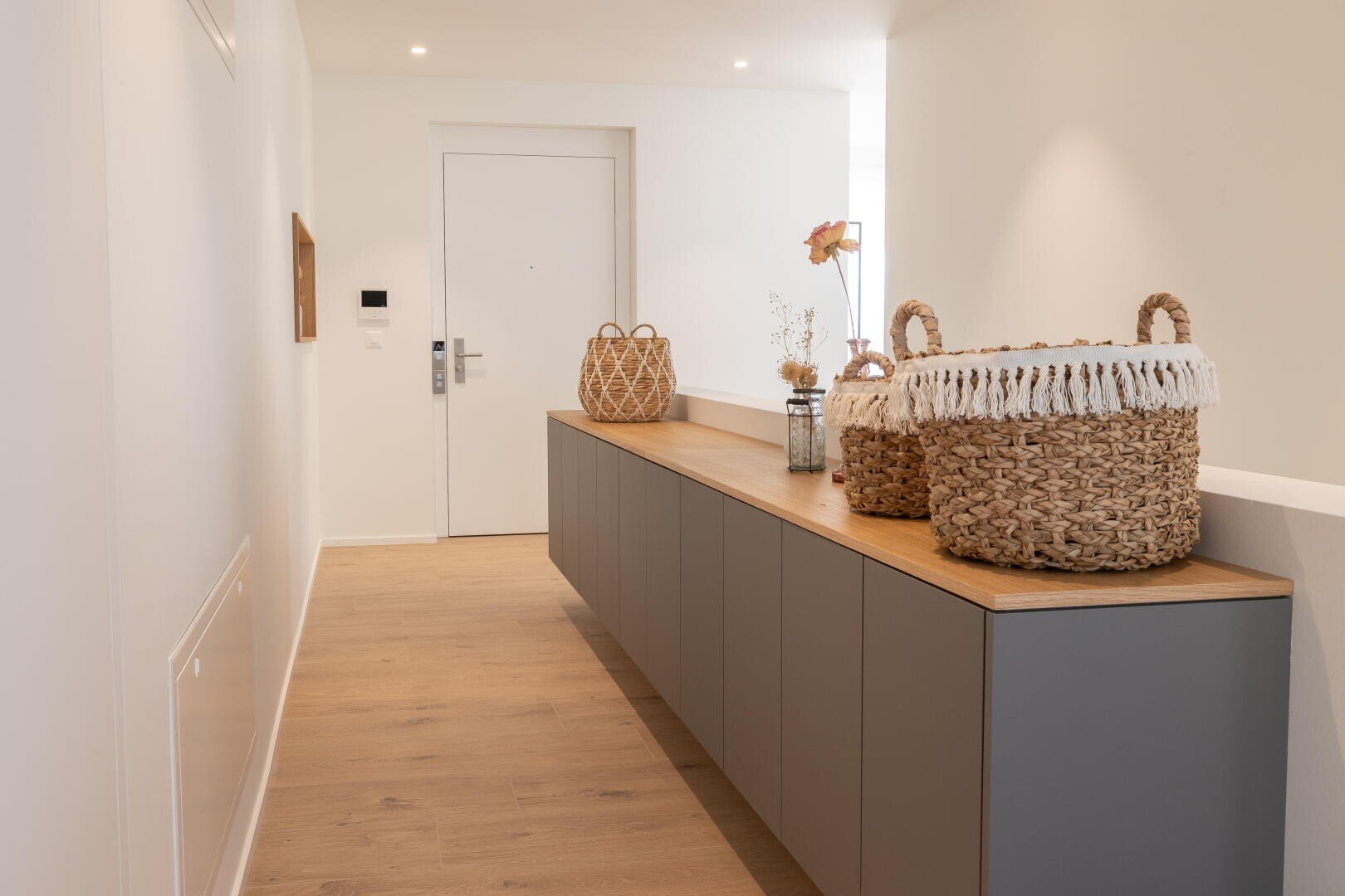 Ein moderner Flur mit hellem Holzfußboden, grauen Schränken mit einer Holzoberfläche und dekorativen geflochtenen Körben und Vasen. Am Ende befindet sich eine weiße Tür, und der Raum ist hell mit neutralen Tönen.