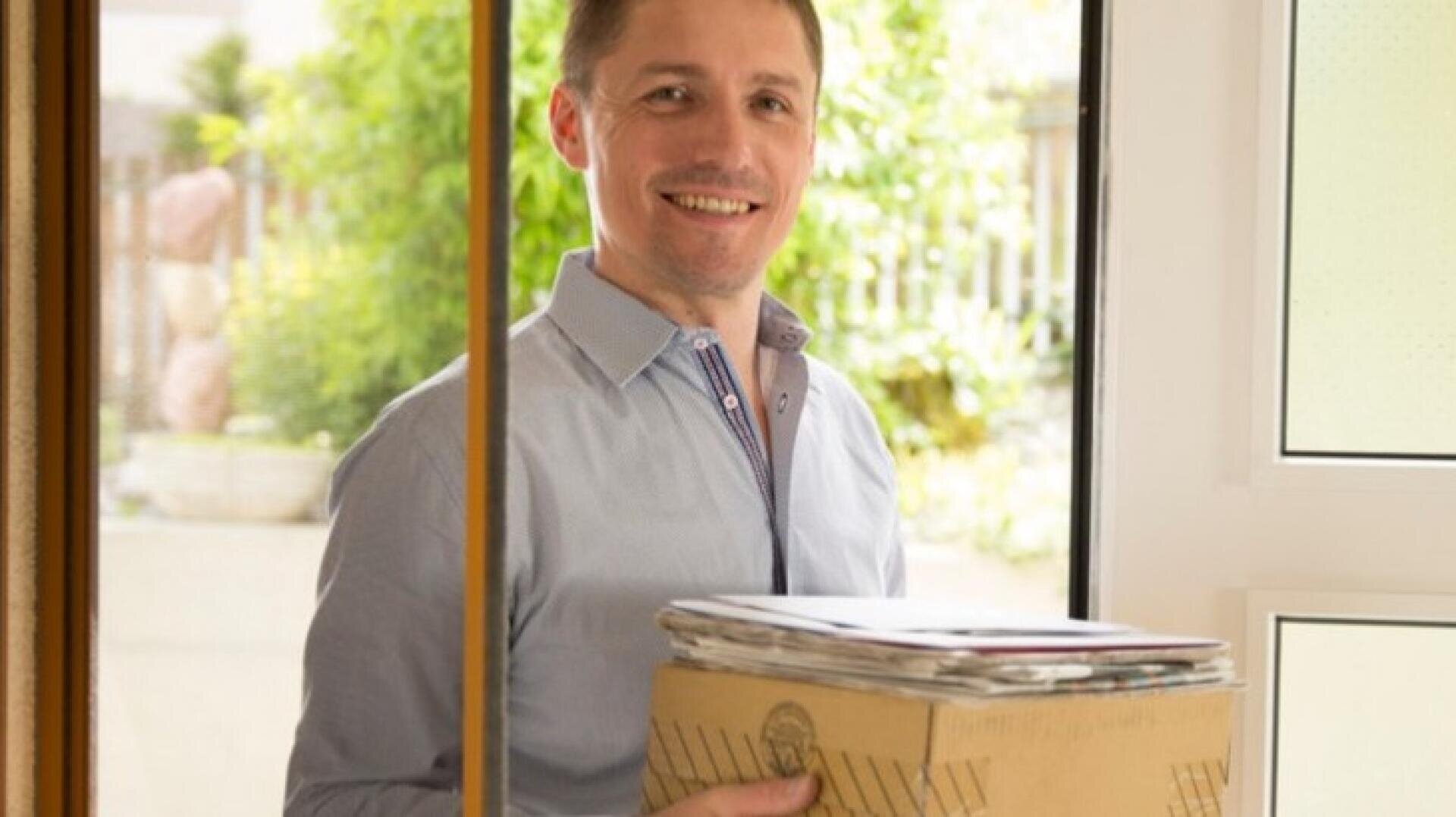 Ein lächelnder Mann in einem hellblauen Hemd steht an einer Tür und hält einen Karton mit Papieren und Zeitschriften in der Hand. Durch das Fenster ist draußen Grünzeug zu sehen.