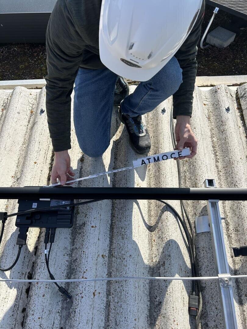 Eine Person mit weißem Schutzhelm und Jeans hockt auf einem Wellblechdach und installiert oder prüft Geräte mit der Aufschrift ATMOC, wobei Kabel und elektronische Geräte in der Nähe sichtbar sind.