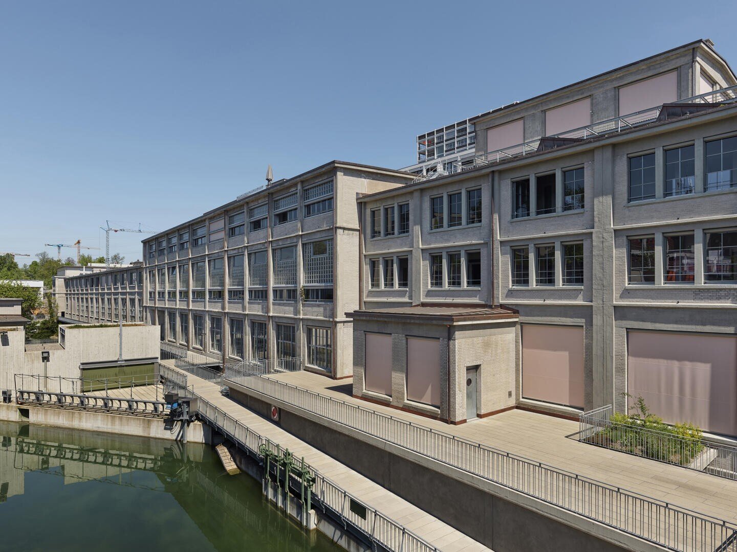 Ein großes, modernes Industriegebäude mit vielen Fenstern und hellen Wänden steht an einem Gewässer unter einem klaren blauen Himmel. Balkone und Gehwege verlaufen entlang der Gebäudekante.