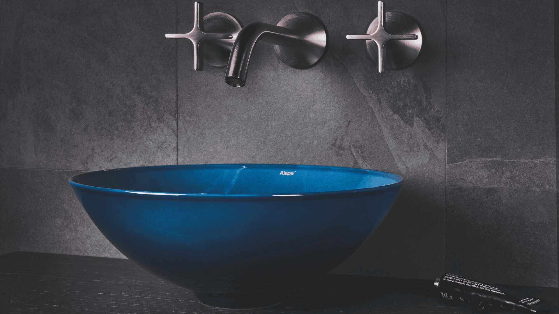 Ein modernes Badezimmerwaschbecken besteht aus einem dunkelblauen Waschbecken und einer Wandarmatur aus gebürstetem Metall mit X-förmigen Griffen, alles auf dunkelgrauen Steinfliesen.