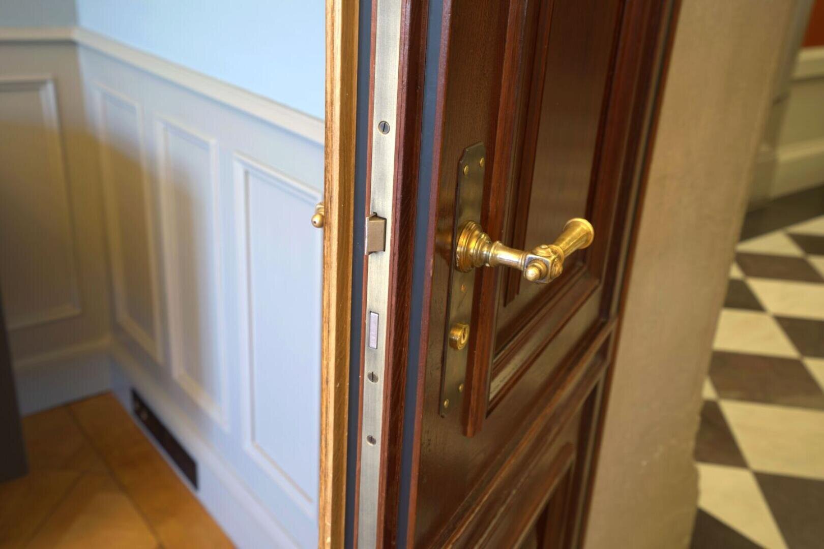 Une porte en bois avec une poignée en laiton est partiellement ouverte et donne sur une pièce avec des lambris bleu clair et un sol avec un motif en damier noir et blanc.