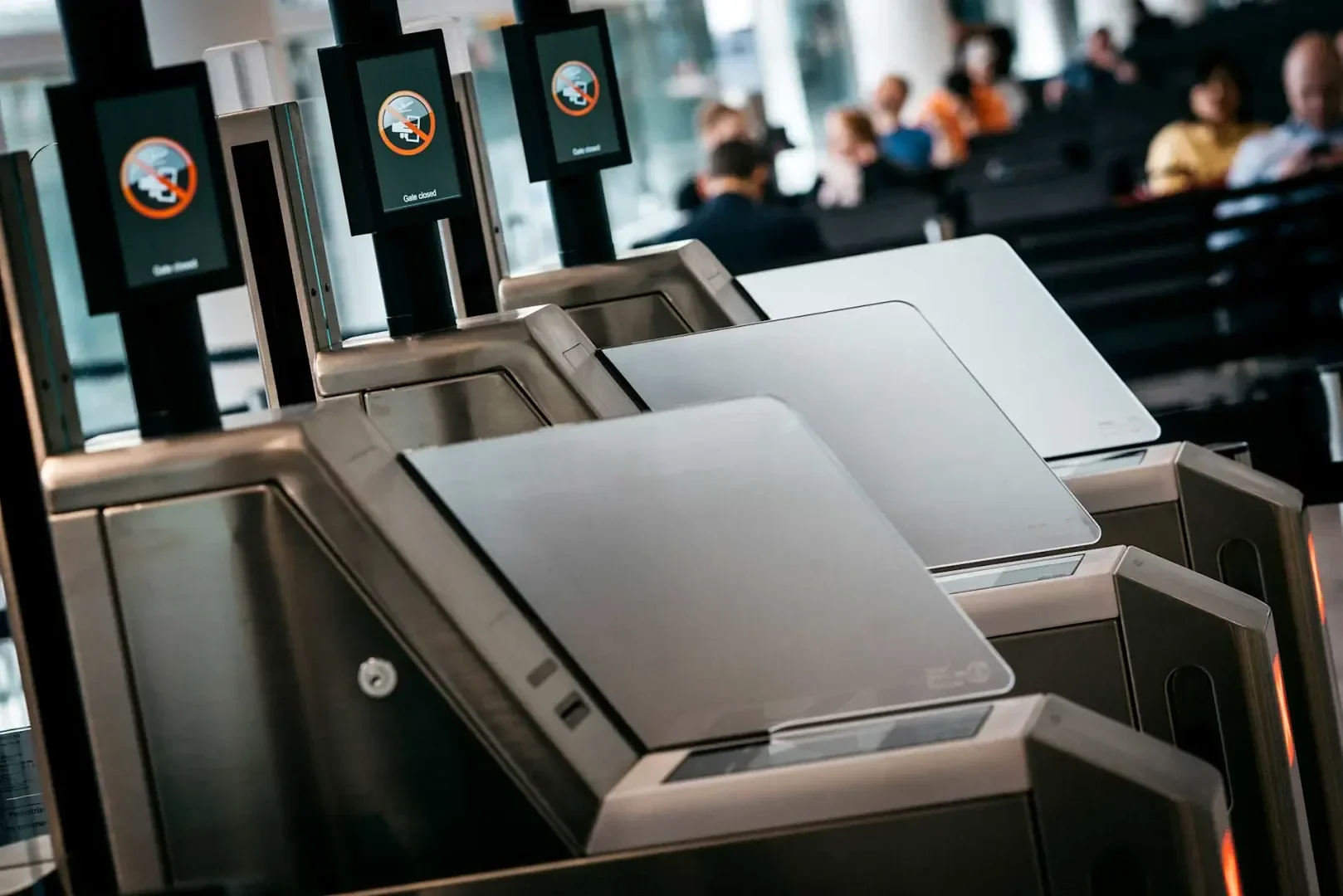 Selbstbedienungs-Check-in-Automaten an einem Flughafen mit digitalen Bildschirmen, auf denen ein "Zutritt verboten"-Schild angezeigt wird. Im Hintergrund sitzen und warten Menschen in einem Terminalbereich.