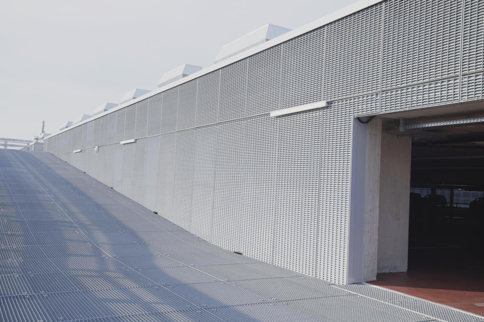 Un bâtiment industriel moderne avec une façade en métal tissé et une rampe menant à une entrée ouverte. La structure se caractérise par des lignes épurées et des motifs géométriques sous une lumière naturelle claire.