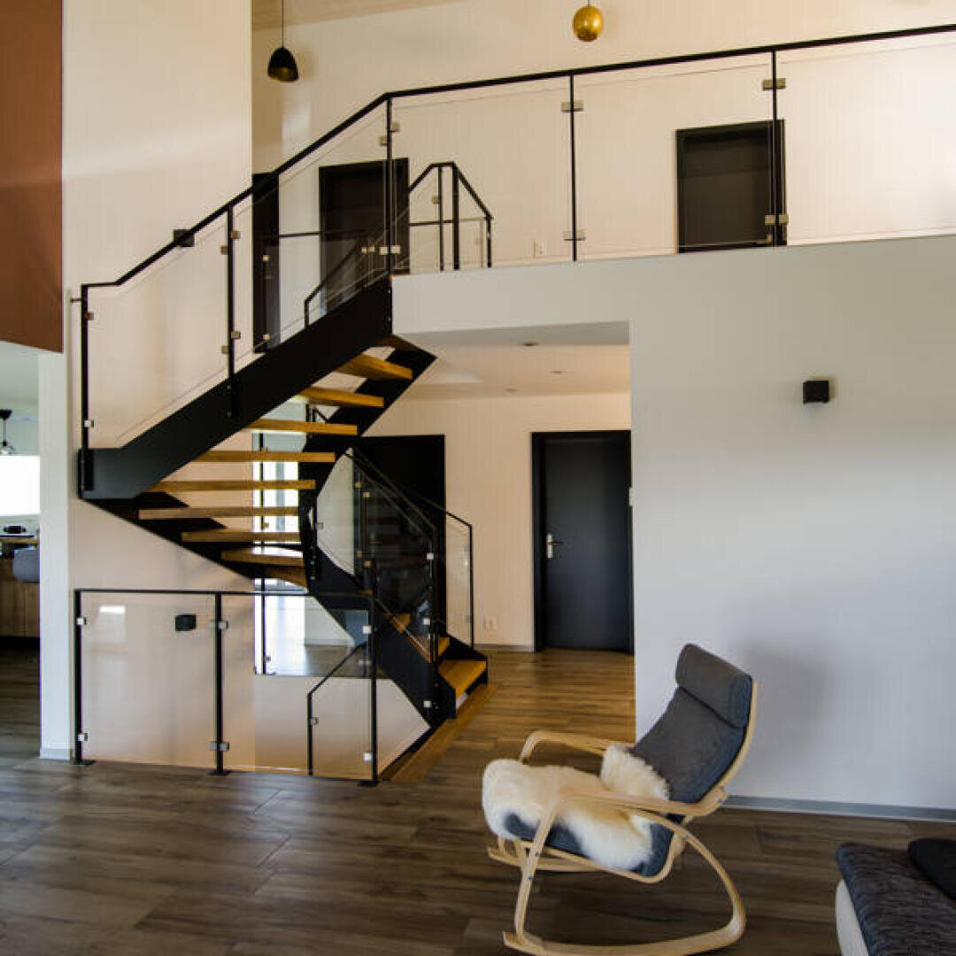 Intérieur moderne avec un sol en bois, un escalier noir sobre avec une rampe en verre, des murs blancs et un fauteuil à bascule recouvert de fourrure près de l'escalier.