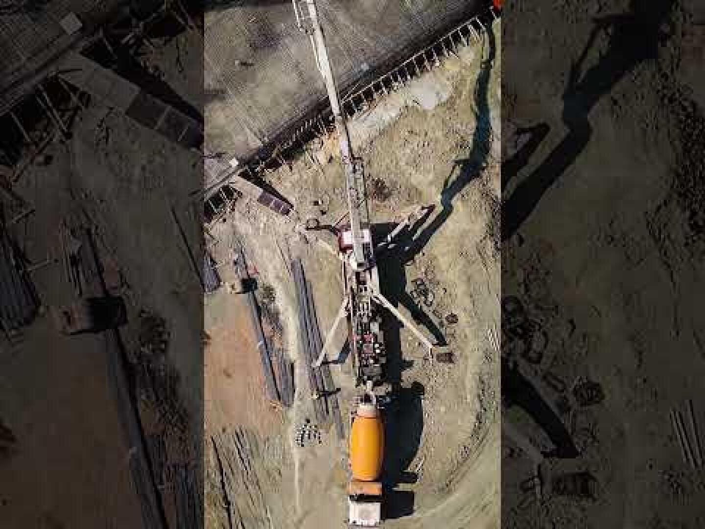 Luftaufnahme einer Baustelle mit einem Betonmischer und einem langen Betonpumpenarm, der zum Gießen von Beton ausgefahren wird, umgeben von Baumaterialien und Schmutz.