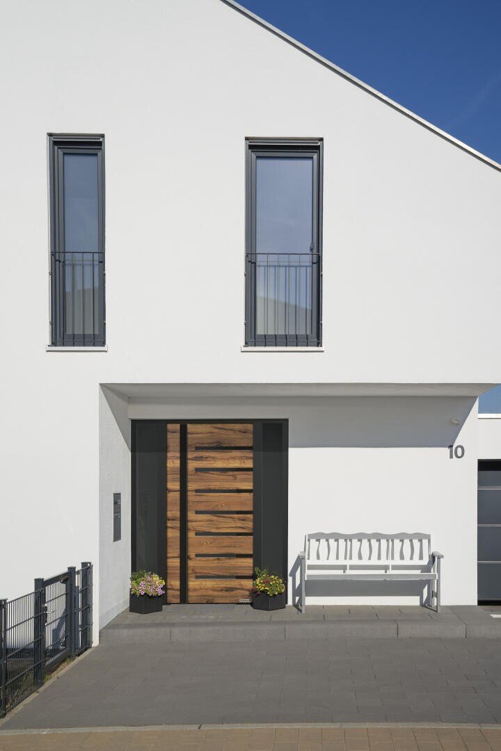 Moderne weiße Hausfassade mit zwei hohen Fenstern über einer hölzernen Eingangstür. In der Nähe des Eingangs befinden sich eine weiße Bank und zwei Topfpflanzen, und an der Wand ist die Hausnummer 10 zu sehen.