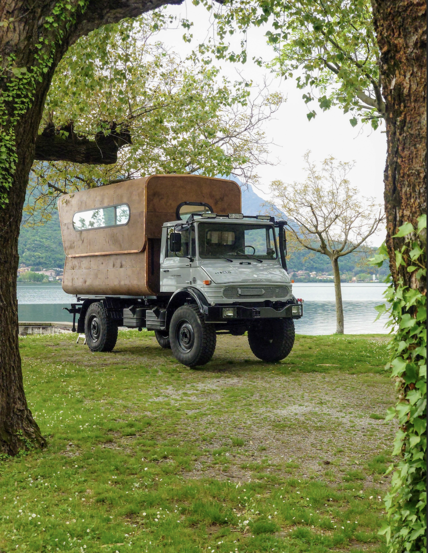 Ein robuster Geländewagen mit einem großen, braunen Wohnmobil ist auf grünem Gras in der Nähe eines Sees geparkt, umrahmt von belaubten Bäumen und Bergen im Hintergrund.