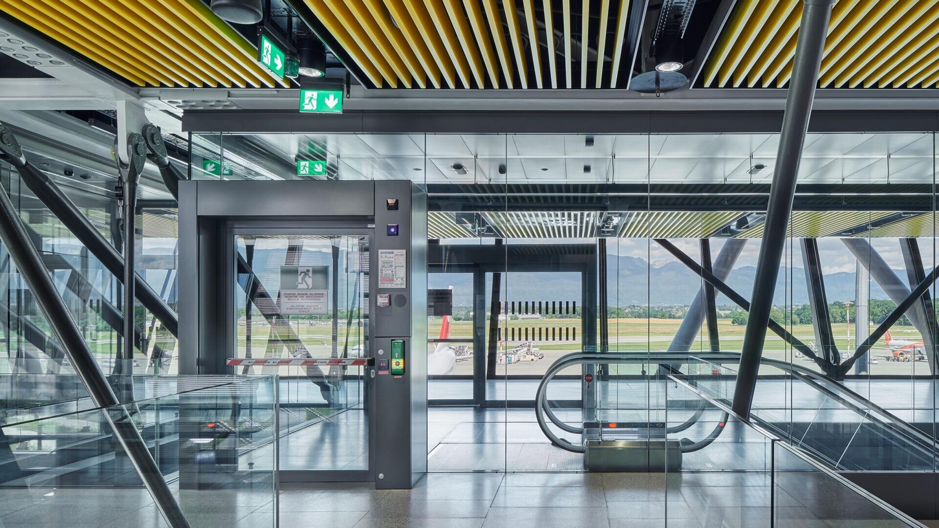 Ein modernes Flughafenterminal mit Glaswänden, einem Aufzug und einer Rolltreppe. An der Decke sind grüne Ausgangsschilder zu sehen, und von draußen hat man einen Blick auf die Landebahn und die Berge.