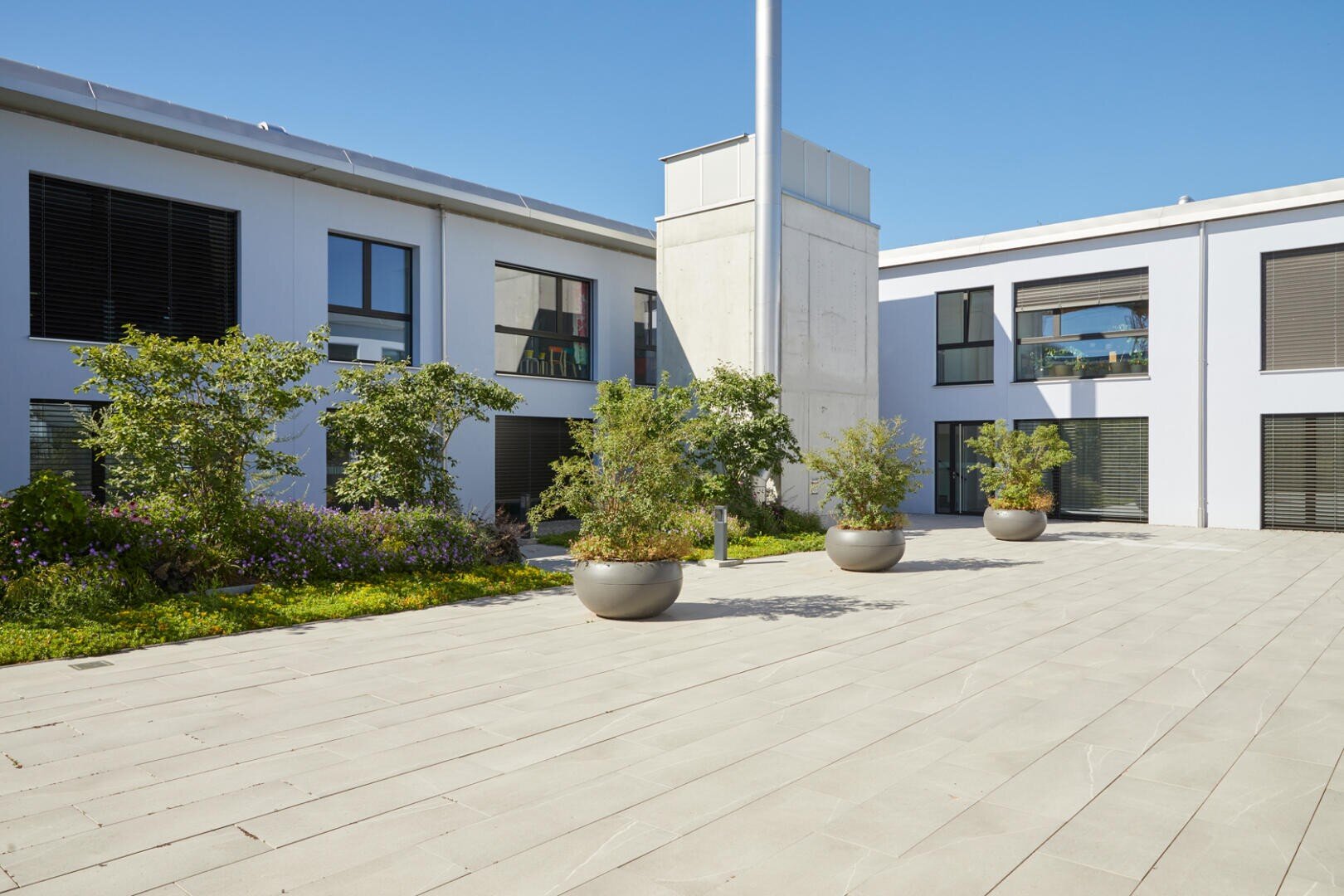 Un bâtiment moderne avec de grandes fenêtres et une cour intérieure avec des jardinières de petits arbres et d'arbustes devant un ciel bleu éclatant.