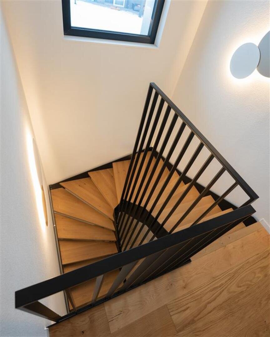Un escalier intérieur moderne en colimaçon avec des marches en bois et une rampe en métal noir, vu d'en haut. Une fenêtre et des appliques murales rondes éclairent l'espace aux murs blancs.