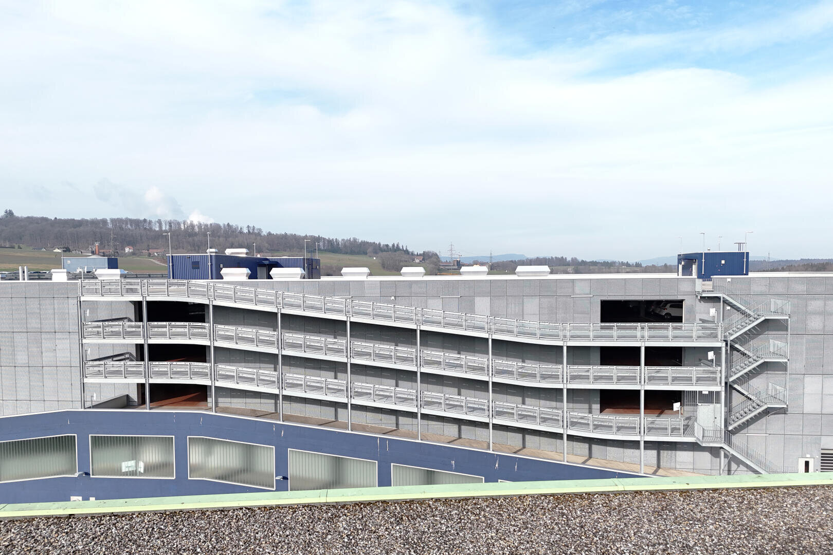 Un parking à plusieurs étages à l'extérieur gris et aux rampes en zigzag, entouré d'un paysage ouvert et d'un ciel partiellement nuageux en arrière-plan.