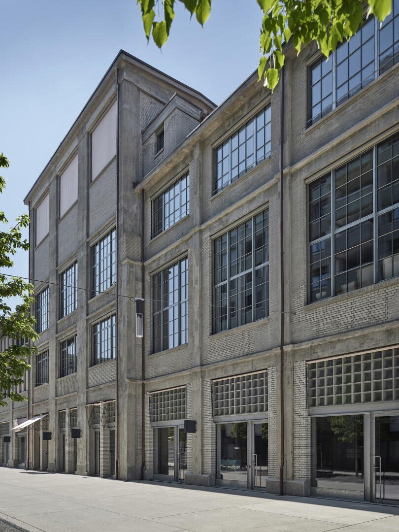 Ein großes Gebäude im Industriestil mit hohen, rechteckigen Fenstern und strukturierten Betonwänden vor einem klaren blauen Himmel. Grüne Baumzweige rahmen die Szene in den oberen Ecken ein.